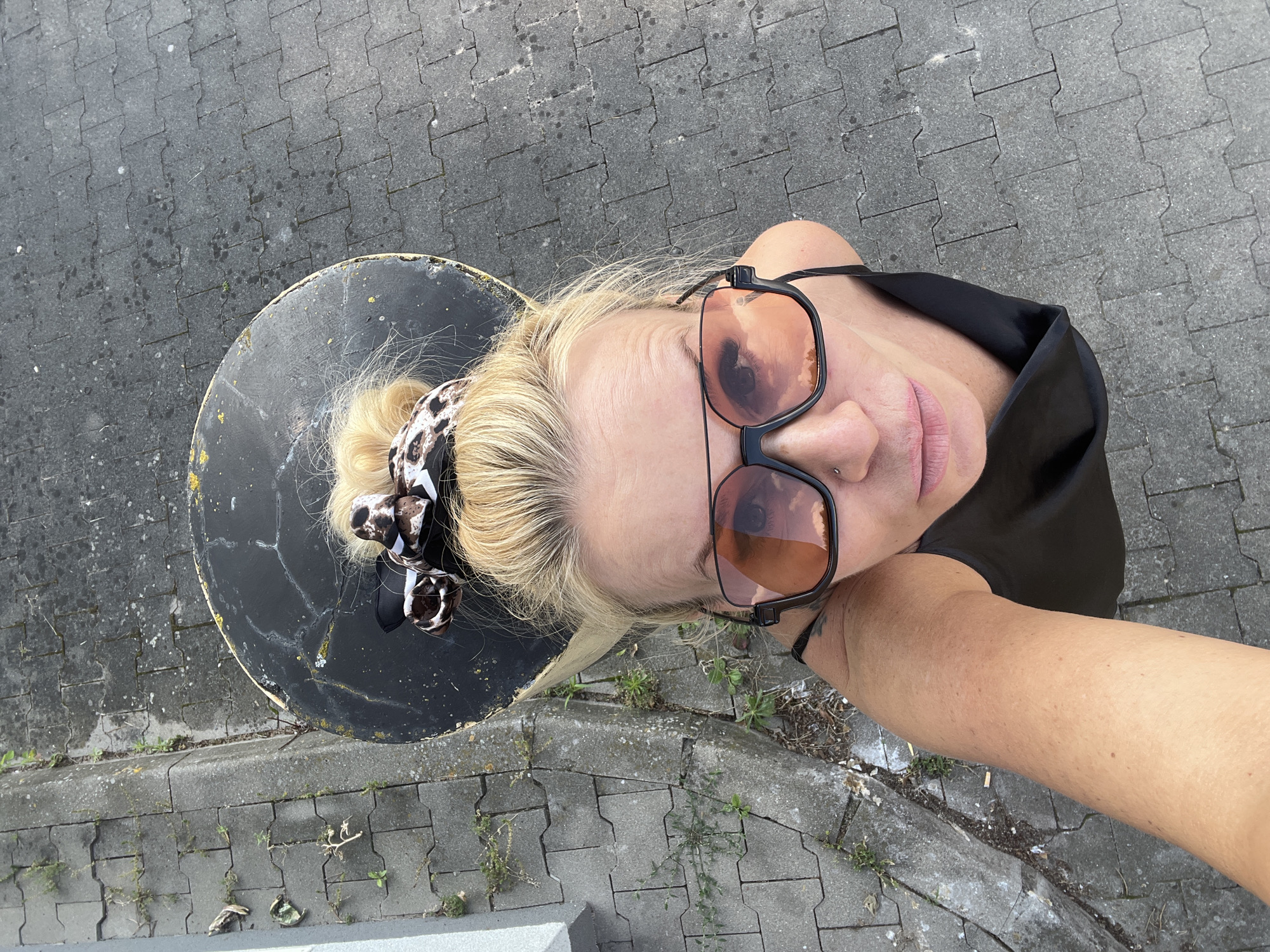 The image shows a woman from a high-angle perspective, looking up at the camera. She has blonde hair pulled up in a bun with a leopard-print scarf. She is wearing large, dark-rimmed sunglasses with orange lenses and a black tank top. Her left arm is extended, holding the camera for a selfie. The background consists of gray paver stones with some patches of dirt and small weeds growing between them. To the left of the paver stones is a concrete curb and a portion of a light-colored wall. In the upper center of the image is a large, black, cylindrical object with a worn surface and yellowing edges. The overall scene appears to be an outdoor urban environment, possibly near a street or sidewalk.