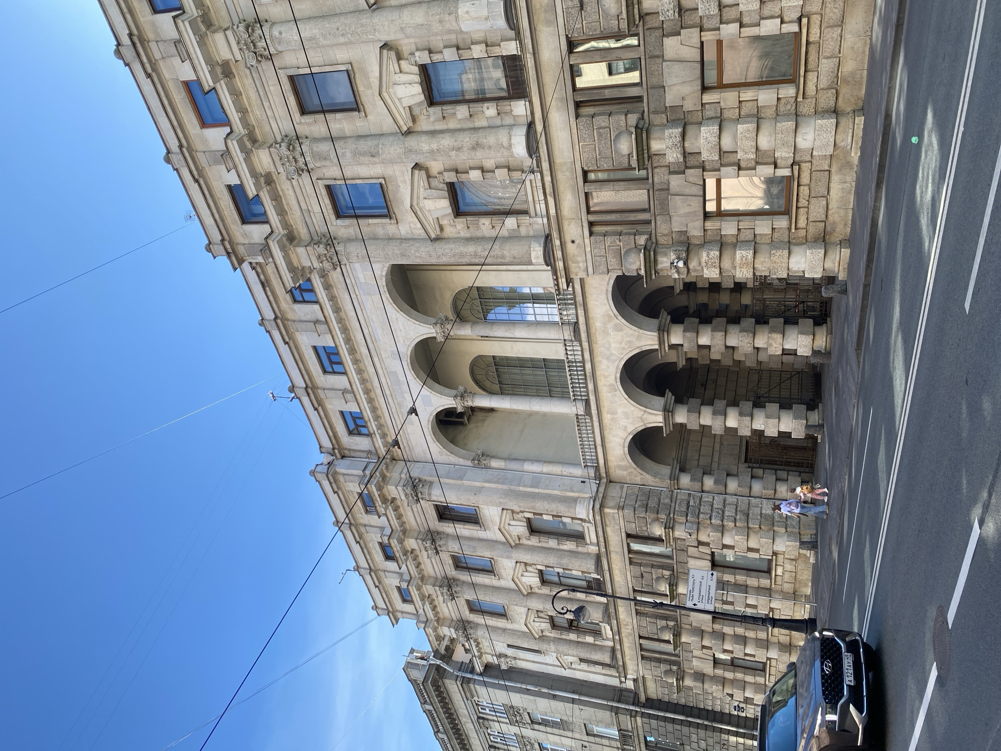 The image displays a sunny daytime street scene in Saint Petersburg, Russia, featuring a grand architectural building alongside a city street. The large, multi-story edifice on the left exhibits classical design elements, including light-colored stone, ornate columns, arched windows and doorways on lower levels, and decorative cornices. A section of the building to the right features distinct rusticated stonework. Overhead, numerous trolley or power lines crisscross the clear blue sky.
On the right, a paved road with white double solid and single dashed lane markings is visible. A dark-colored Hyundai SUV, bearing the Russian license plate "A121AT 98", is parked or moving on the street. Two individuals are discernible on the sidewalk in front of the building, near a black street lamp. A street sign, written in Cyrillic, points to several local streets, including "Большого проспекта П.С. 7", "Барочная улица, 5", "Ул. Подрезова, 13", "Сытнинская улица, 12", and "Кронверкская улица, 11", confirming the location context. Bright sunlight illuminates the scene, casting shadows on the road and highlighting architectural details. A small green object rests on the roadway.