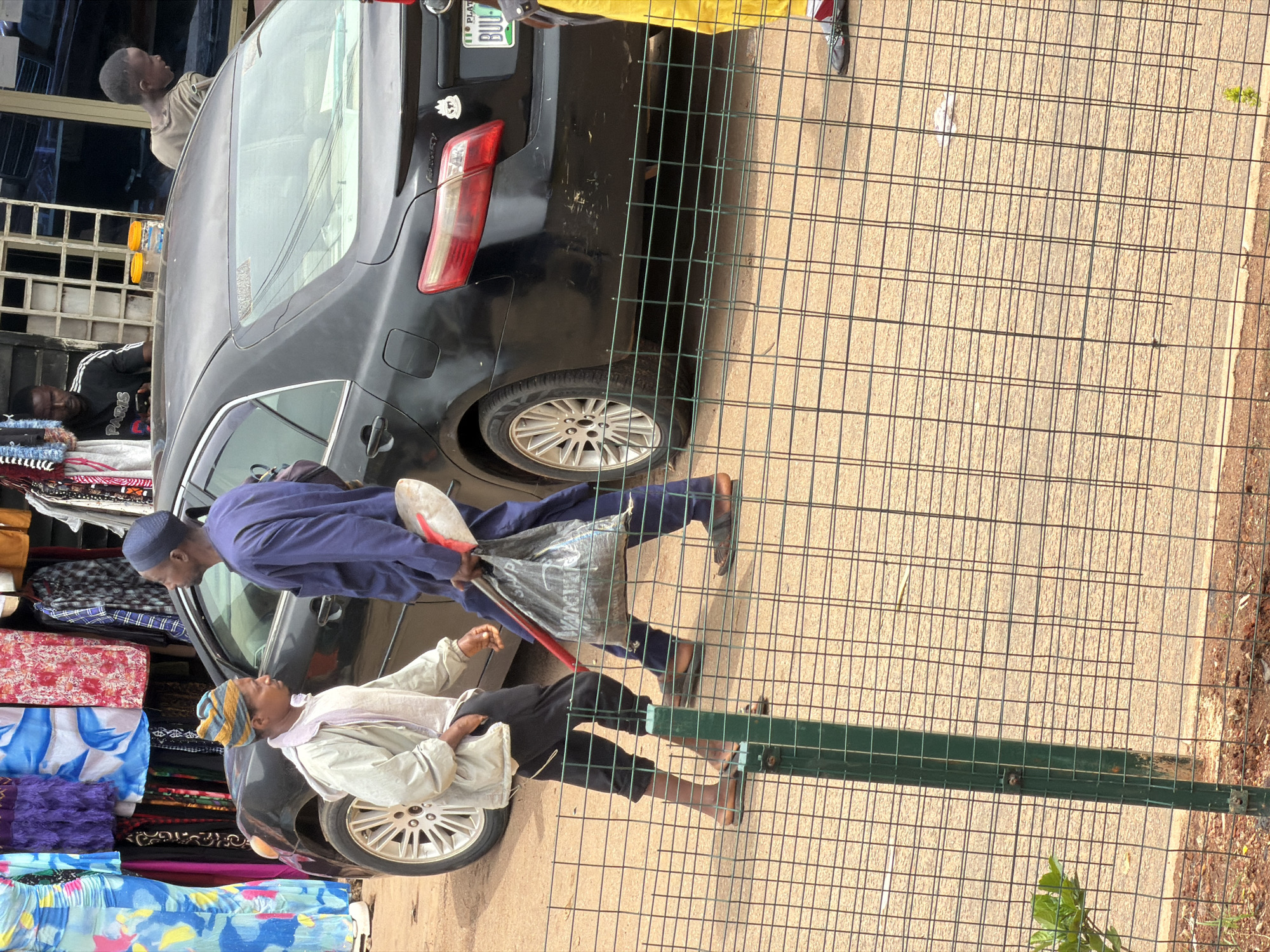 In Bukuru, Nigeria, two men are walking next to a parked black sedan. The man on the left wears a light-colored jacket and dark pants, with a striped beanie. The man on the right is dressed in a blue tunic and trousers and carries a black plastic bag containing a red-handled shovel. The men are walking on a dirt road, and a wire fence is visible in the foreground. In the background, there is a stall displaying colorful fabrics, and other individuals are visible near a building. The license plate of the car reads "PLAT BUU-".