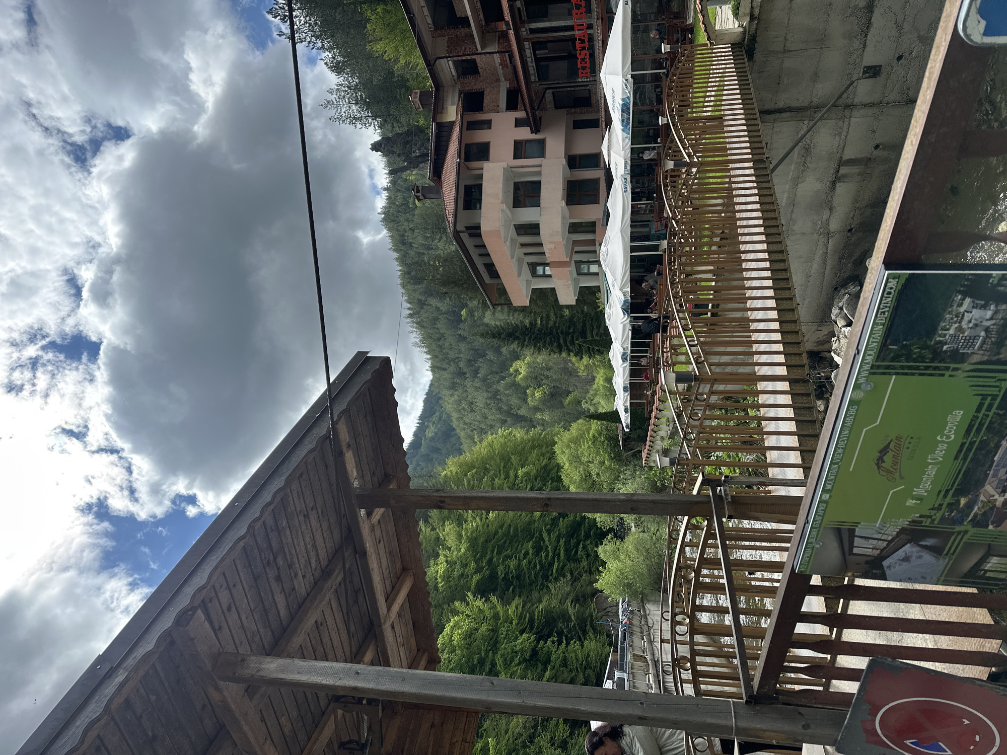 The image shows an outdoor scene in Iagodina, Bulgaria. A partially visible wooden roof structure is in the foreground, with a dense forest on a hillside in the midground. Adjacent to the roof structure is a wooden fence with a metal railing. Behind the fence, there is a building with multiple stories, colored in pastel shades, and a restaurant with outdoor seating and umbrellas. The sky is filled with dark, cloudy skies, with some patches of blue visible. A sign with the text "Mountain View Ecovilla" is partially visible on the right side of the frame. A bridge or walkway appears to be constructed over a stream or river. The angle of the photo causes the objects in the scene to appear tilted.