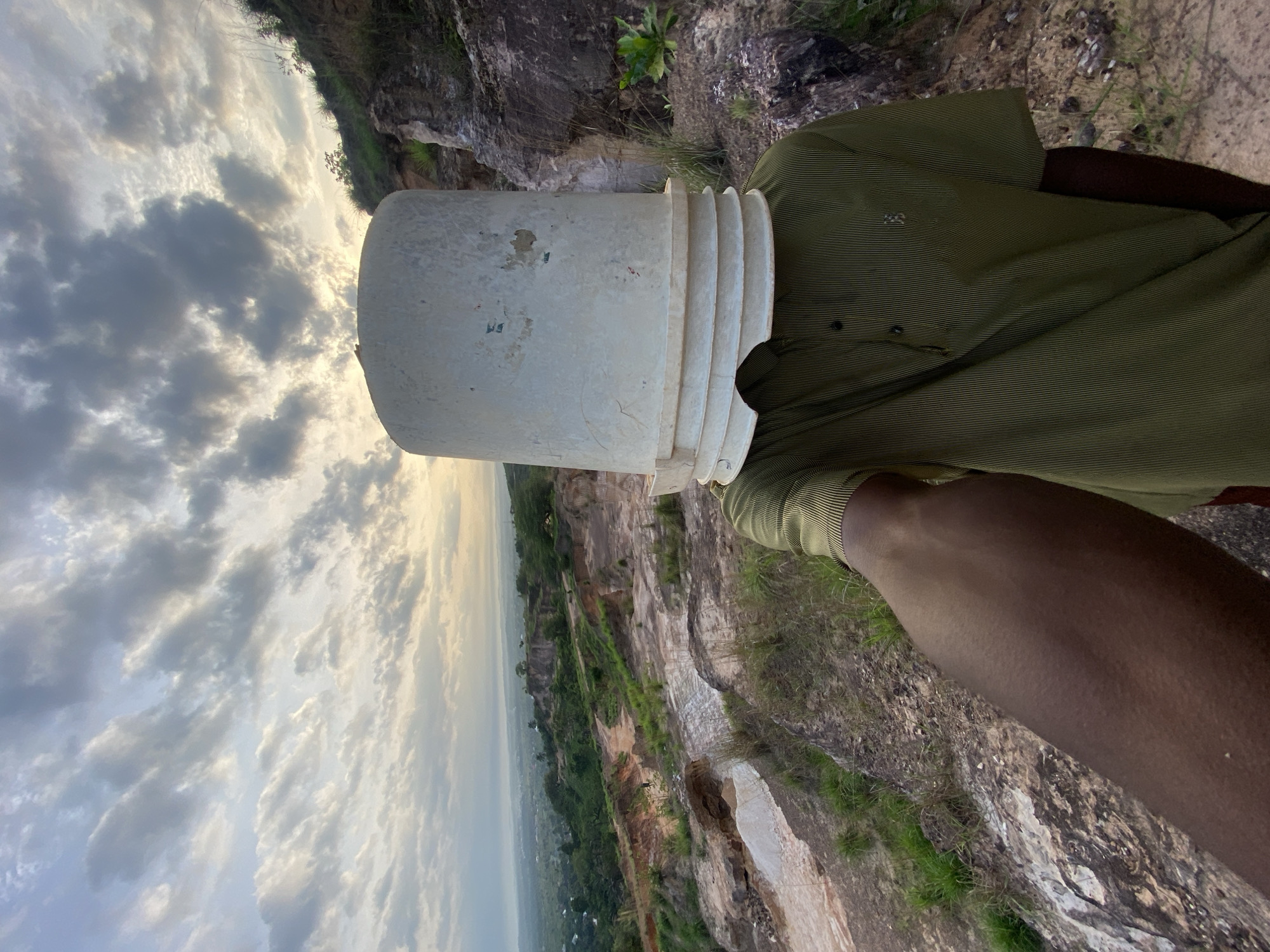 An outdoor, eye-level shot captures a person standing on a rocky outcrop overlooking a vast landscape. The individual, whose skin appears dark, is seen from the chest down, wearing an olive green shirt with subtle horizontal stripes and black buttons. Their head is entirely obscured by a large, white, somewhat scuffed plastic bucket, which has several ridged layers near its rim.
The person is positioned near the edge of what appears to be a large quarry or a rugged, elevated terrain with extensive exposed rock formations and steep, bare slopes. Patches of green vegetation cling to the rocky surfaces, indicating some plant life amidst the harsh environment. The background reveals a broad valley stretching into the distance, with hazy contours of distant hills or settlements under a partly cloudy sky. The lighting suggests either early morning or late afternoon, with a soft, diffused light illuminating the scene and casting a gentle, slightly golden glow on the clouds. The sky is filled with a mix of white and darker grey clouds, indicating partly cloudy conditions.
No specific activity beyond standing and observing is discernible, and with the head covered by the bucket, no emotions are visible from the person. Faint marks and possibly faded text are present on the bucket, but no writing is clearly legible. A small, indistinct emblem is visible on the left chest area of the person's shirt. The scene conveys a sense of scale and natural grandeur within the location of Ufueseni, Nigeria.