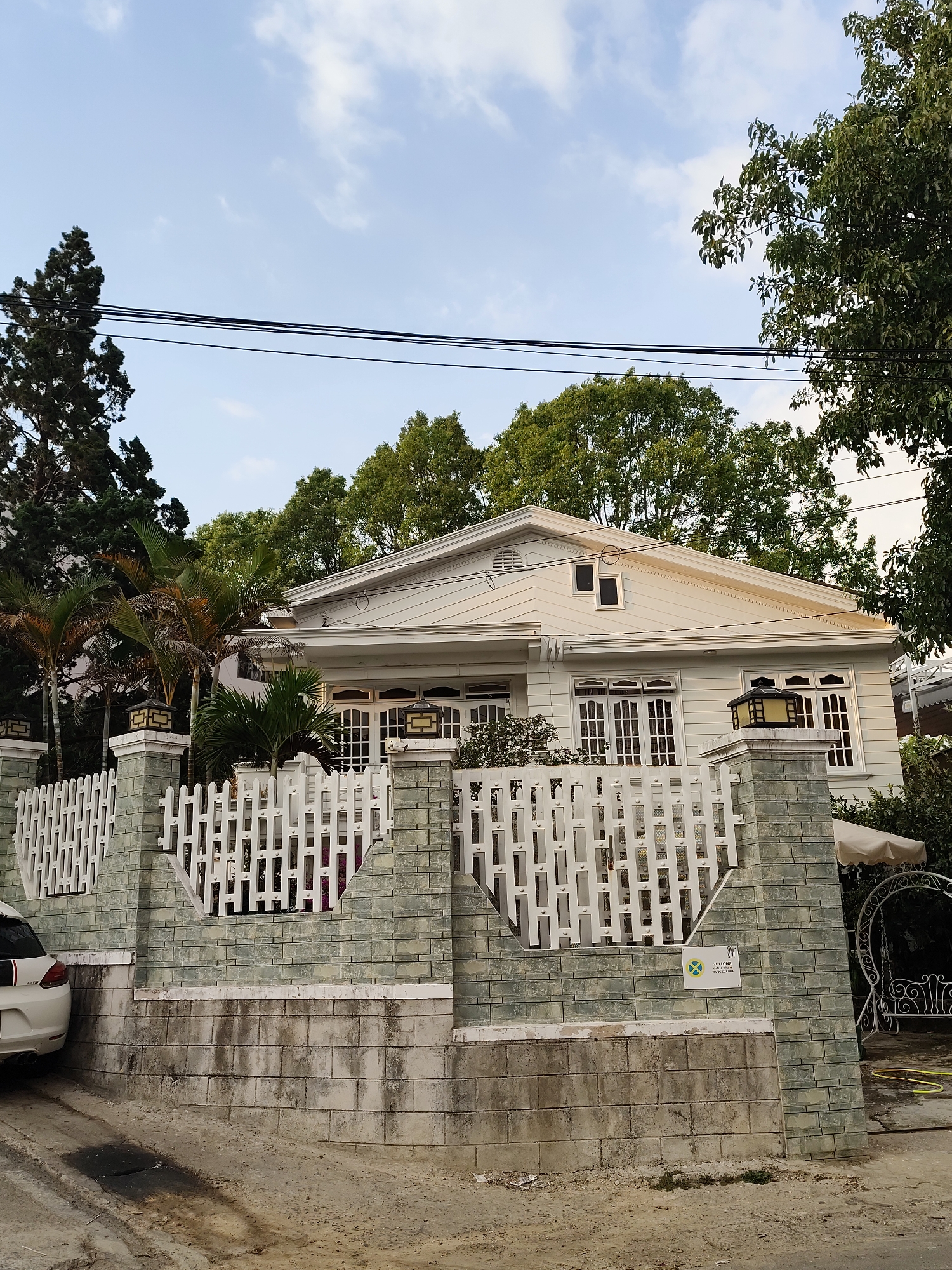 A daytime exterior shot reveals a white, single-story house with a triangular gable roof, observed behind a low stone and white picket fence. The house features multiple windows with white frames, a small attic window centered in the gable, and decorative lighting fixtures mounted on the fence posts. The fence consists of gray-green stone pillars connected by white, vertical picket panels. A partial view of a white car is visible to the left of the fence. Lush green trees are prominent in the background, partially obscuring the view of the sky, which shows a clear gradient of blue. Power lines stretch across the upper portion of the frame. A small sign is affixed to the stone fence. The ground in front of the fence appears to be an unpaved street or pathway. The location is suggested to be Da Lat, Vietnam.
