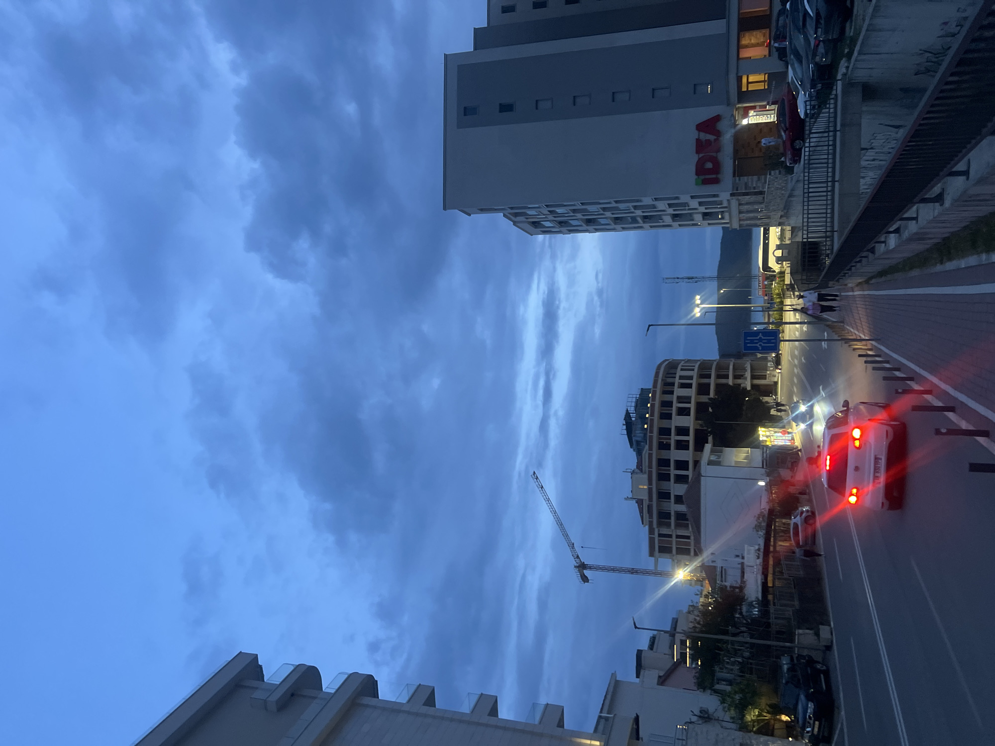 The image shows a street scene in Boreti, Montenegro at dusk. The sky is overcast with blue clouds. On the right side of the image, a white car with illuminated red taillights is stopped on a two-lane road. A sidewalk runs along the right edge of the road, and a metal railing separates the sidewalk from an elevated area.
On the left side of the image, several buildings of different heights are visible. One building has the word "IDEA" in large red letters. In the middle of the image, there is a construction crane. The building architecture is modern. The overall lighting suggests early evening.