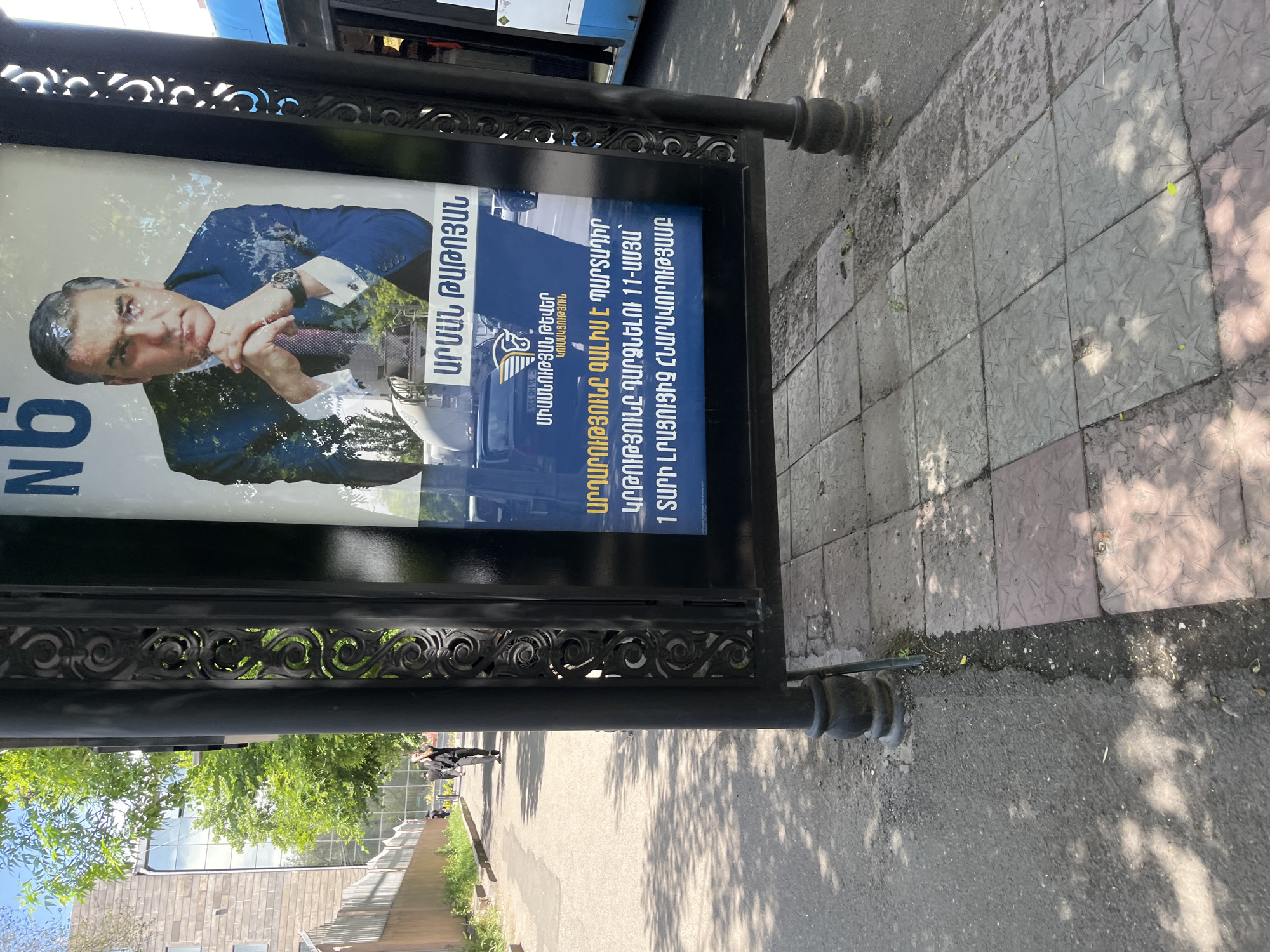 Here's a detailed description of the image:
The image captures a political poster displayed on a black, ornate stand at a street level, likely in Yerevan, Armenia. The poster features a man in a suit, with text in Armenian. Some of the text reads "ԱՐՄԱՆ ԹԱԹՈՅԱՆ" and mentions education and justice. The poster appears to be an advertisement for a political candidate.
The environment includes a sidewalk composed of grey and pink square tiles with star pattern, with a street visible beyond, along with a bus. There are also trees and a building visible in the background. Shadows from trees and sunlight fall across the scene.