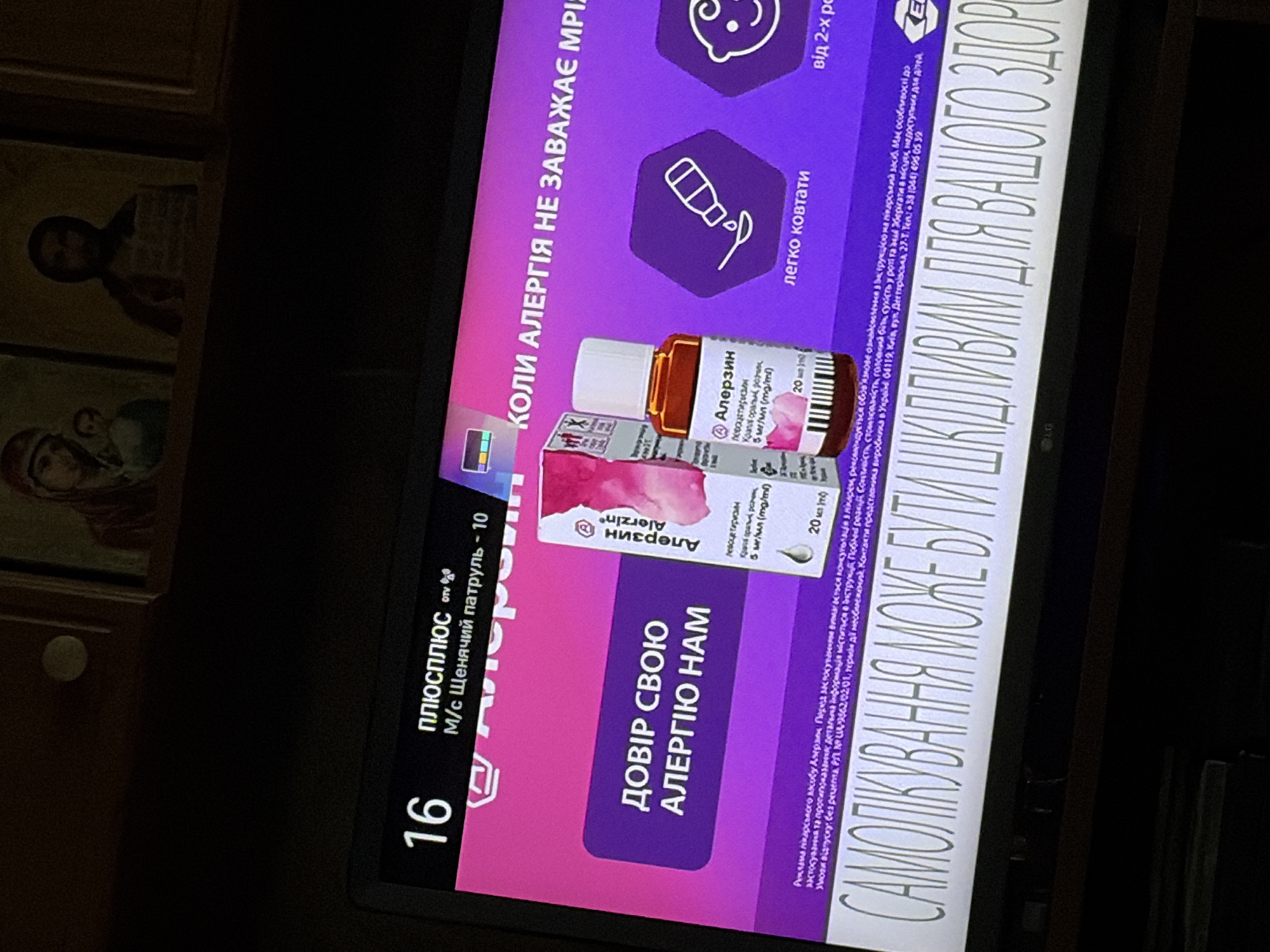 A television screen displays an advertisement for an allergy medication called "Алерзин" (Alerzin). The ad features a pink and purple color scheme. On the left side, the number "16" is prominently displayed. Below it, the text reads "плюсплюс" and "M/с Щенячий патруль - 10". A box and a small bottle of the medication are shown. The text "ДОВІР СВОЮ АЛЕРГІЮ НАМ" (Trust your allergy to us) is written in large white letters on a purple background. To the right, there are icons representing a spoon with liquid and a smiling baby. The slogan "КОЛИ АЛЕРГІЯ НЕ ЗАВАЖАЄ МРІЯМ" (When allergy doesn't stop your dreams) is written in Ukrainian. At the bottom of the screen, a large tagline in Ukrainian reads "САМОЛІКУВАННЯ МОЖЕ БУТИ ШКІДЛИВИМ ДЛЯ ВАШОГО ЗДОРОВ'Я" (Self-medication can be harmful to your health). Below this tagline, smaller text provides important disclaimers and contact information. The background appears to be a dimly lit room with framed religious icons visible above the television screen.