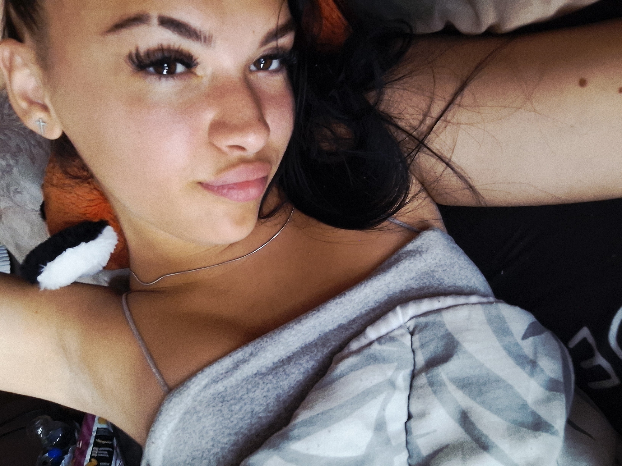 A young adult female is depicted lying on her back, looking towards the camera with her head tilted to the right. She has dark hair, brown eyes, prominent dark eyelashes, and full, light-pink lips. A small silver-toned cross-shaped earring is visible on her left ear, and a thin silver necklace rests around her neck. She is wearing a light gray camisole with spaghetti straps. A patterned blanket with light gray stripes covers her lower torso and left arm. Her right arm is visible, displaying several small moles on the skin. Partially visible behind her head is a reddish-brown stuffed animal with a white and black ear. The background shows blurred domestic items, including a plastic water bottle. The scene takes place indoors. Based on the visual content provided, there is no information to establish the location as Krapets, Bulgaria.