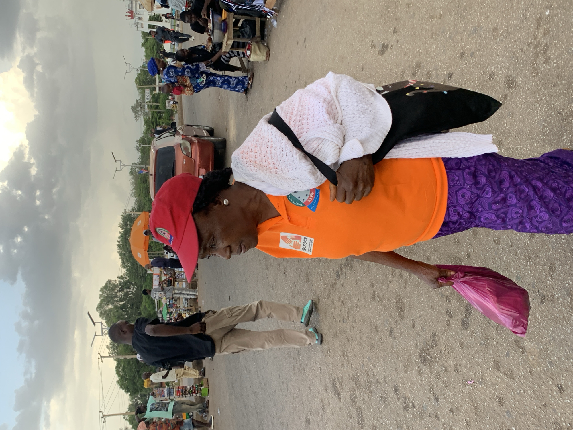 In a bustling urban environment, a woman in an orange shirt and purple skirt walks on a paved road in Jos, Nigeria. She wears a red cap and a white knitted shawl draped over her shoulders. A black bag hangs on her shoulder, and she carries a pink plastic bag in her right hand. The background reveals a busy street with several people, vehicles, and market stalls. The sky is overcast with clouds. The scene captures a moment of everyday life in a Nigerian city, showcasing the vibrant atmosphere of the local community.