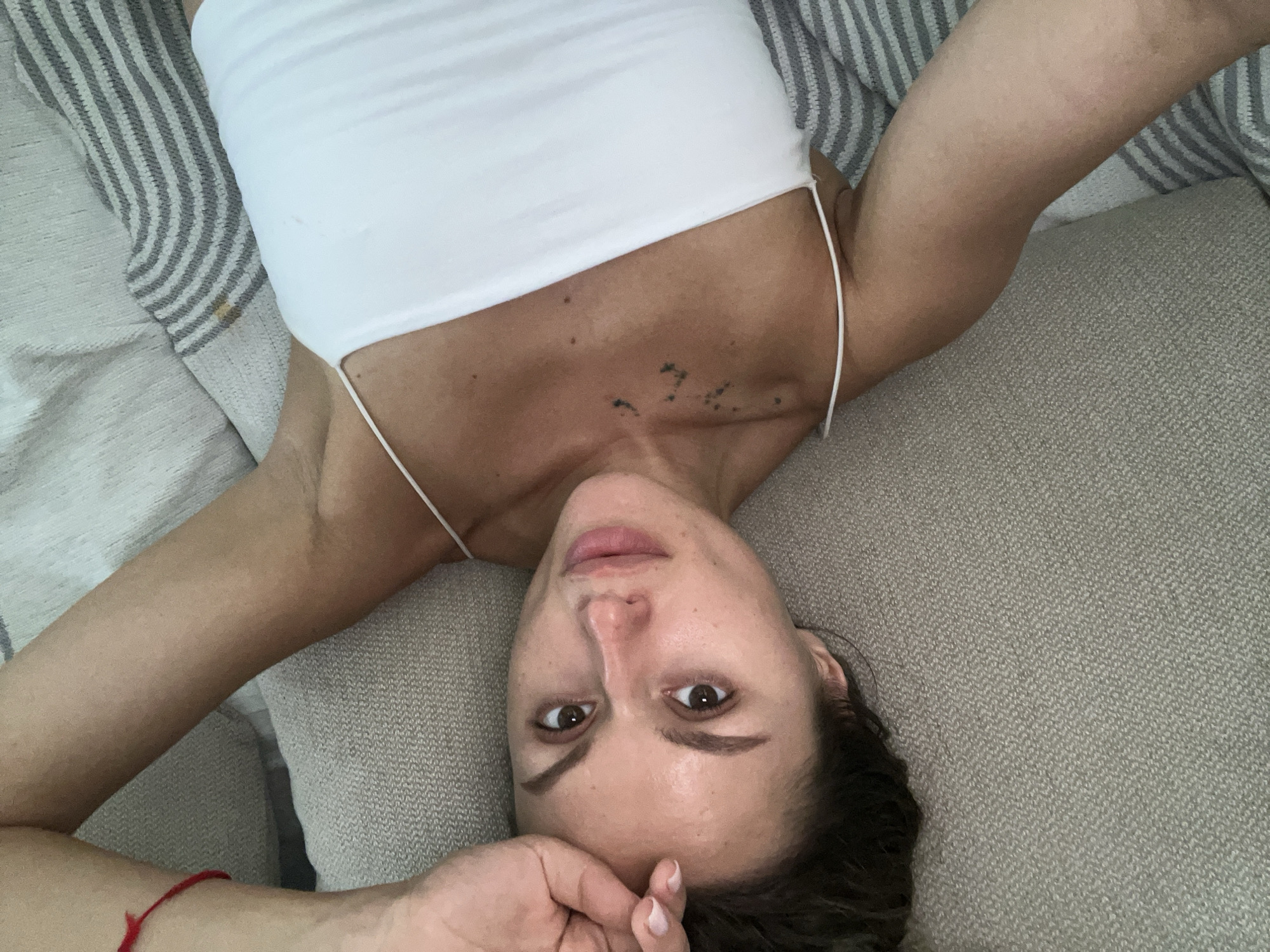 A woman lies on her back on a textured grey surface, likely a couch or bed. She is wearing a white camisole. Her right arm is bent, with her hand resting on her forehead. Her left arm is also bent, with her elbow resting on the surface. The woman has brown hair pulled back, and her face is turned towards the camera. She has brown eyes and light skin. There are faint tattoos on her upper chest. A red string bracelet is visible on her right wrist. The background consists of grey and white striped fabric. The lighting appears to be natural, coming from above. The overall impression is of a casual, relaxed pose in an indoor setting. There are no discernible elements in the image that specifically place the scene in Choeng Thale, Thailand.