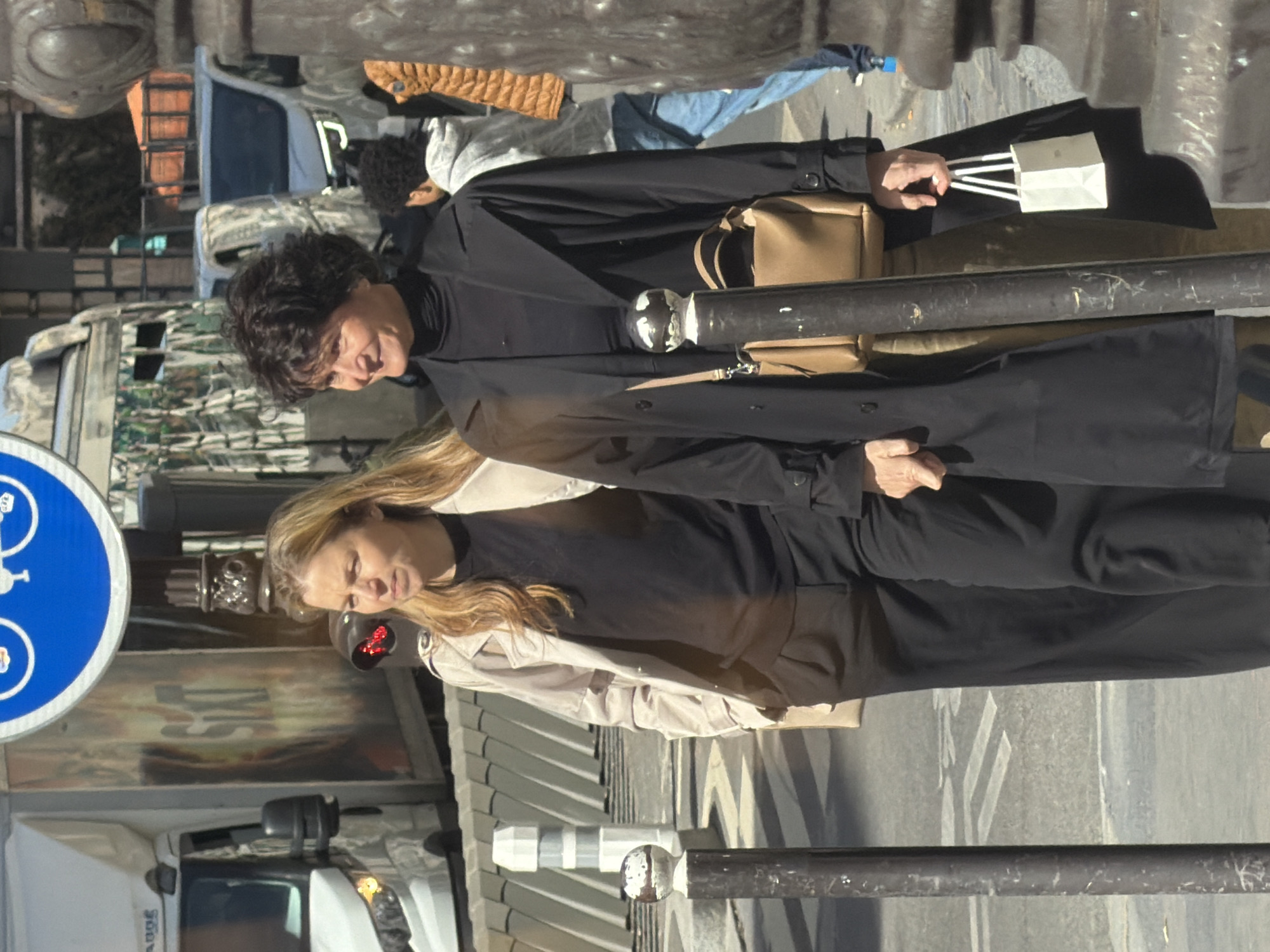 The image captures a Parisian street scene featuring two women. The first woman, with curly dark hair, is clad in a long black coat and carries a small tan handbag. She is also holding a white paper bag with white rope handles. The second woman, with long blonde hair, is wearing a lighter-colored coat over a black shirt. They are standing on a street with pedestrian crosswalk markings, near a metal pole or railing. To the left, a blue circular sign with a bicycle symbol is visible, suggesting a bike lane or designated bicycle area. In the background, there is a white truck and other vehicles, along with hints of buildings. The scene appears to be captured during daylight hours, with a clear sky and bright sunlight casting shadows on the street. The location is likely Paris, France, based on the architectural style and the presence of typical Parisian street signage.