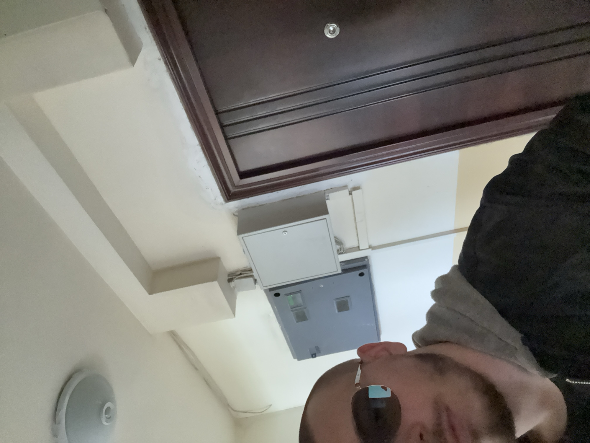 A low-angle shot looking up at a ceiling. The ceiling is white with a dark brown door frame. A circular white light fixture is on the left. A gray electrical box with a white cover is mounted on the ceiling. A person wearing sunglasses, a gray hoodie, and a black jacket is in the foreground. The location is Stara Zagora, Bulgaria.