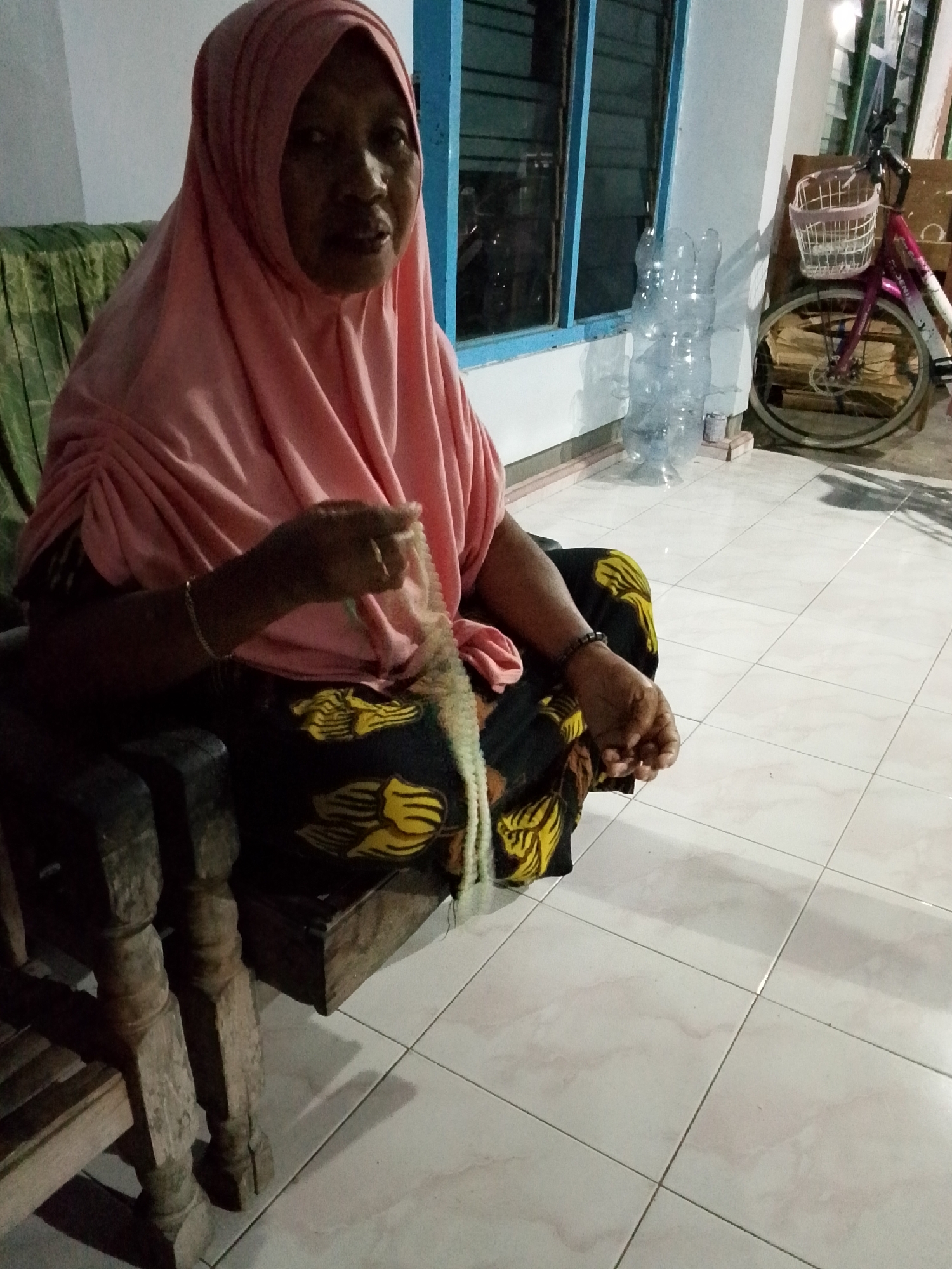 An older woman wearing a pink hijab and a black and yellow patterned sarong sits on a wooden chair. She is holding a green lizard in her right hand. The woman is looking towards the camera. Behind her, a window with blue trim is visible, and to the right, a bicycle with a white basket is parked. The scene takes place on a tiled floor, likely outdoors. The location is identified as Bulurejo, Indonesia.