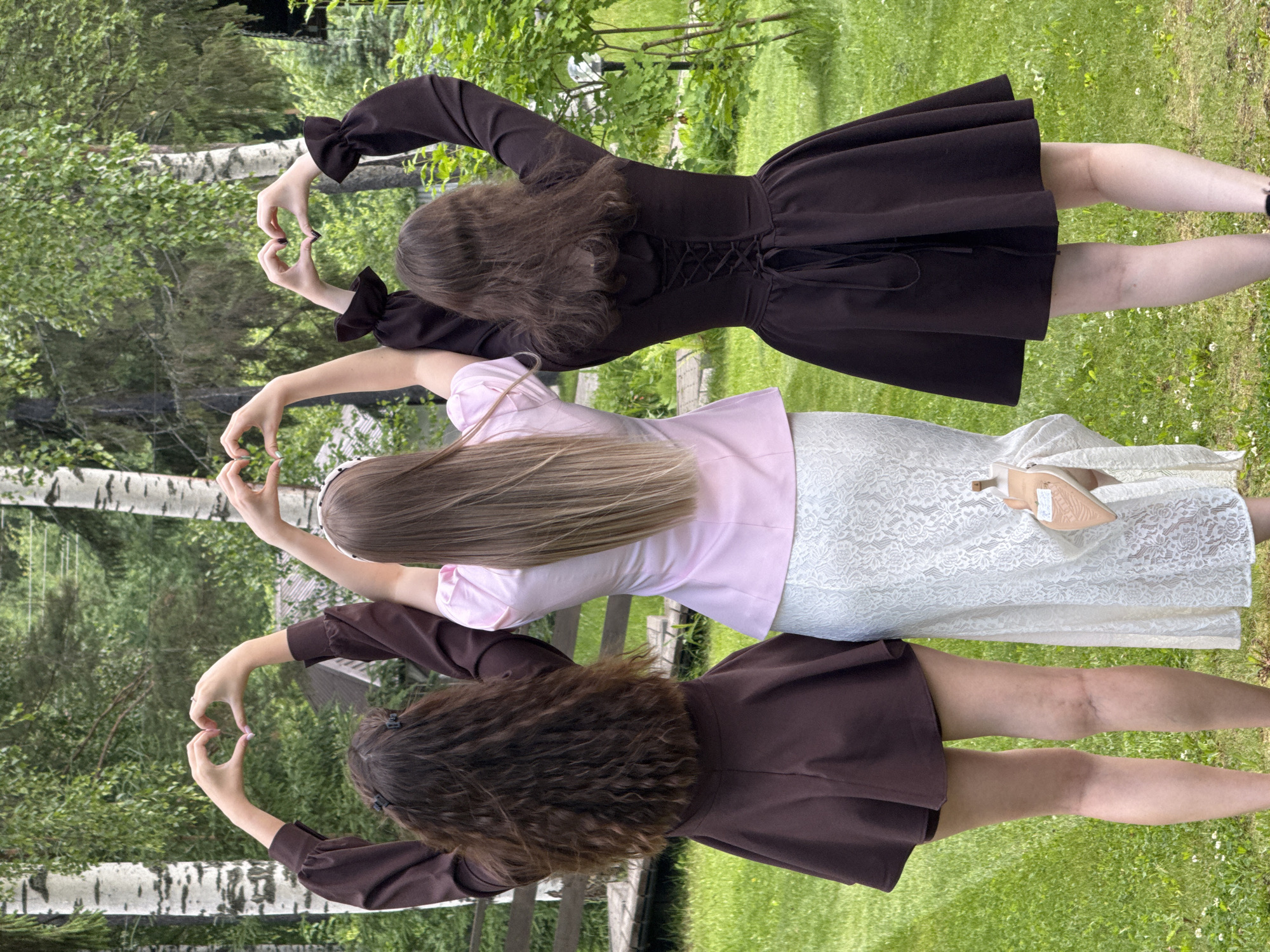 Three individuals are shown from the back, standing outdoors on a grassy area with trees in the background. Each individual is forming a heart shape with their hands above their heads. The person on the left wears a dark brown dress with flared sleeves and a flared skirt. The middle person wears a light pink top and a white lace skirt, holding a tan high-heeled shoe. The person on the right wears a dark brown dress with a corset-style back and a flared skirt. The environment appears to be a natural setting with birch trees and lush green foliage. The scene is captured during daytime, with natural lighting. There is no visible signage or architecture that would definitively place this scene within Votkinsk, Russia.