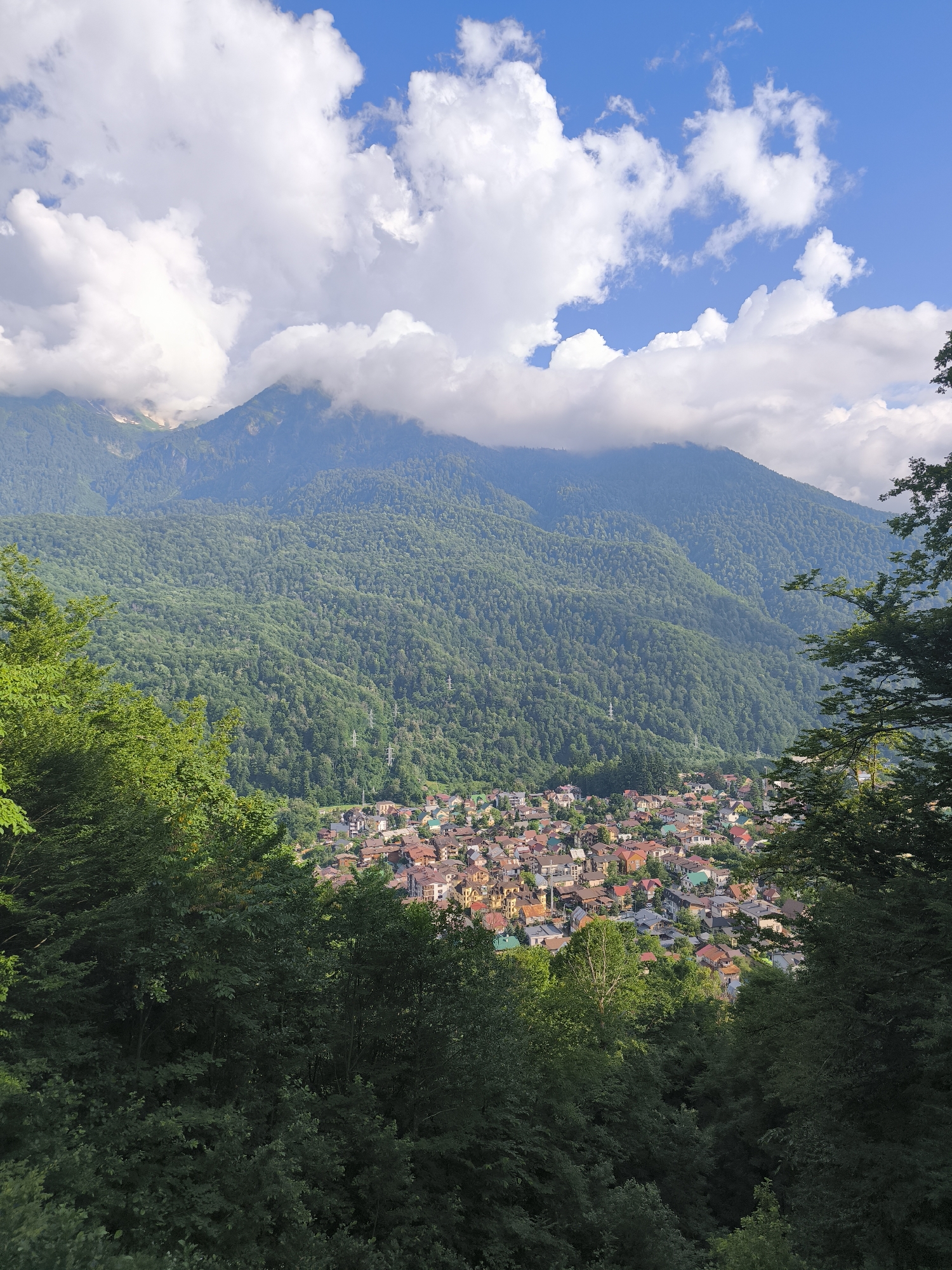 This media file captures a scenic view of a mountain landscape, likely in Krasnaya Polyana, Russia. The scene is dominated by lush green mountains covered in dense forest, with a small village nestled in the valley below. The village features a collection of houses with colorful roofs, creating a vibrant contrast against the natural backdrop.
The sky is a clear blue, dotted with fluffy white clouds, suggesting a pleasant day. The time of day appears to be late morning or early afternoon, as indicated by the bright sunlight casting shadows and illuminating the scene. The weather seems to be fair and warm, ideal for outdoor views.
The foreground is framed by the dark green foliage of trees, adding depth and a sense of perspective to the shot. In the distance, the peaks of the mountains rise majestically, some partially obscured by clouds. Power lines are visible on the slopes of the mountains, indicating human infrastructure within the natural setting.
The overall emotion conveyed by the image is one of tranquility and natural beauty. The juxtaposition of the small, human settlement with the vastness of the mountains evokes a sense of peace and harmony between nature and civilization. There is no visible text in the image.