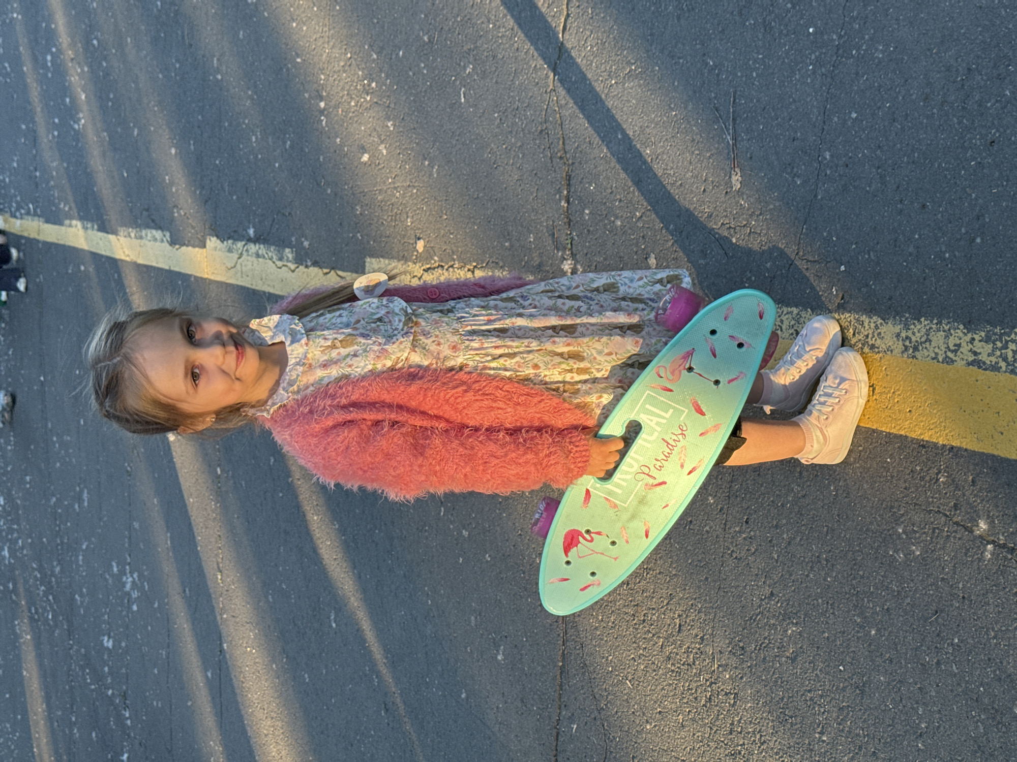 A young girl is lying on a paved surface. She is wearing a floral print dress, a pink fuzzy sweater, and white sneakers. She is holding a teal skateboard with a flamingo design and the words "Tropical Paradise" on it. The paved surface has a yellow line on one side. There are shadows cast across the surface. It is not possible to determine the location of the city of Izhevsk, Russia from the image.
