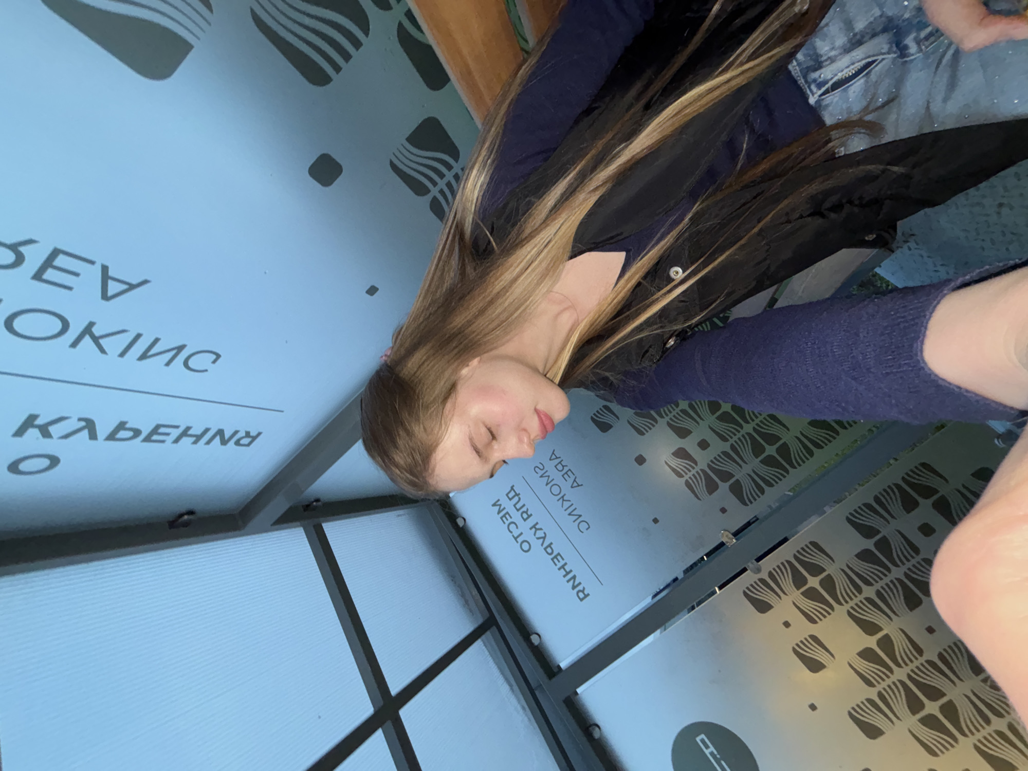 A young woman with long blonde hair is leaning against a wall with her eyes closed. She is wearing a dark blue long-sleeved shirt, a black vest, and sparkly blue jeans. The background features a blue wall with text that appears to be in Russian, along with a smoking area symbol and a decorative pattern. The scene appears to be in a public indoor space, possibly a bus station or waiting area, given the seating and the frosted glass ceiling. The text on the wall is partially visible and includes Cyrillic letters. There is no indication of the location being in the city of Arfa, Russia.