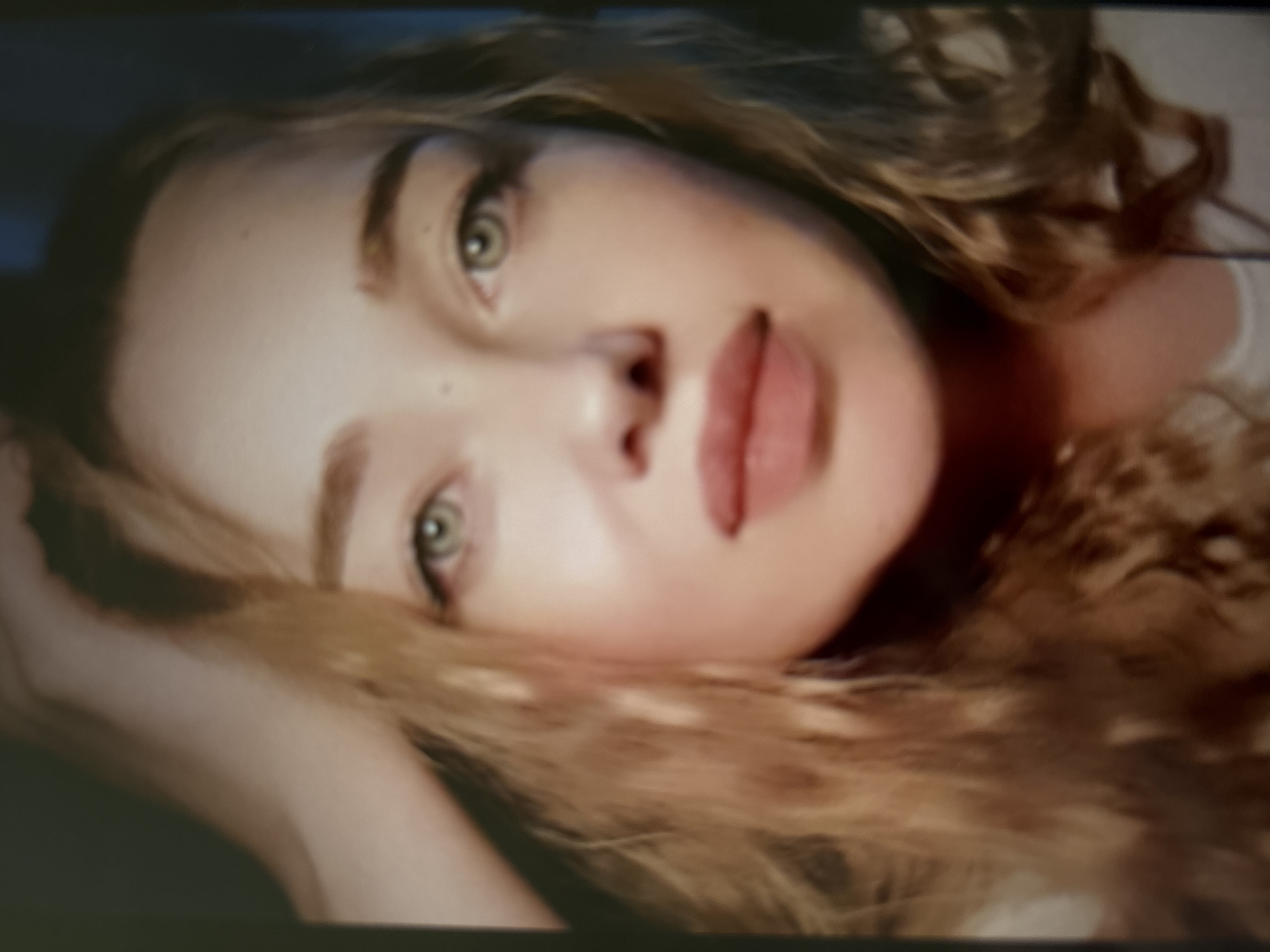 The image is a close-up portrait of a young woman with light-colored, wavy hair. She is looking directly at the camera, with her left hand raised to the side of her head. She is wearing light-colored clothing. The background is dark and out of focus. There is no indication of a specific location, either within Novosibirsk, Russia, or elsewhere.