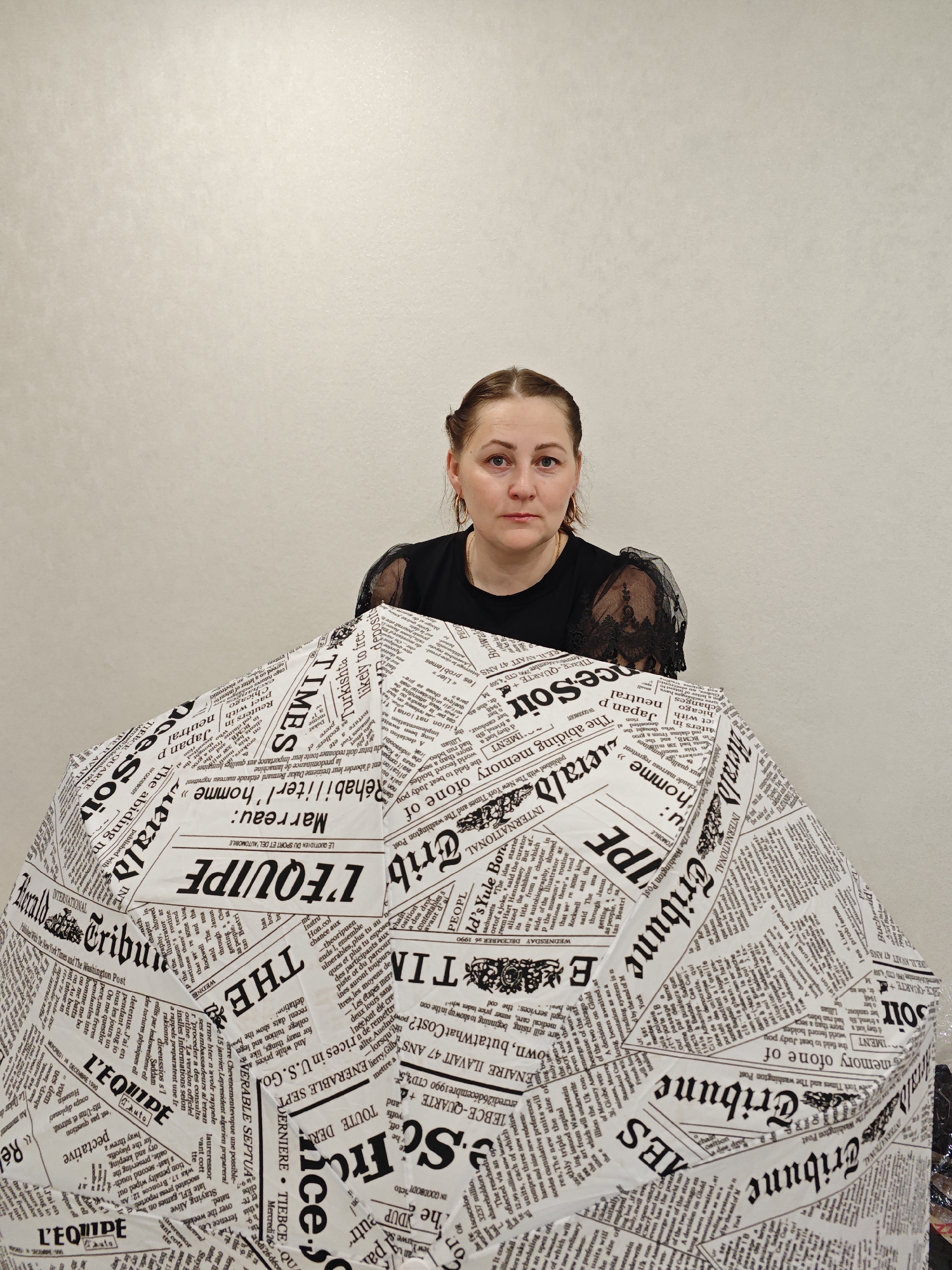 A woman is partially visible behind an umbrella decorated with newspaper print. The woman has brown hair pulled back into a bun and is wearing a black dress with lacy sleeves. She is looking directly at the camera with a neutral expression. The umbrella is covered in text and headlines in black and white. The background is a plain, light-colored wall. The location is likely indoors. The city of Markovskii, Russia is not apparent from the image.