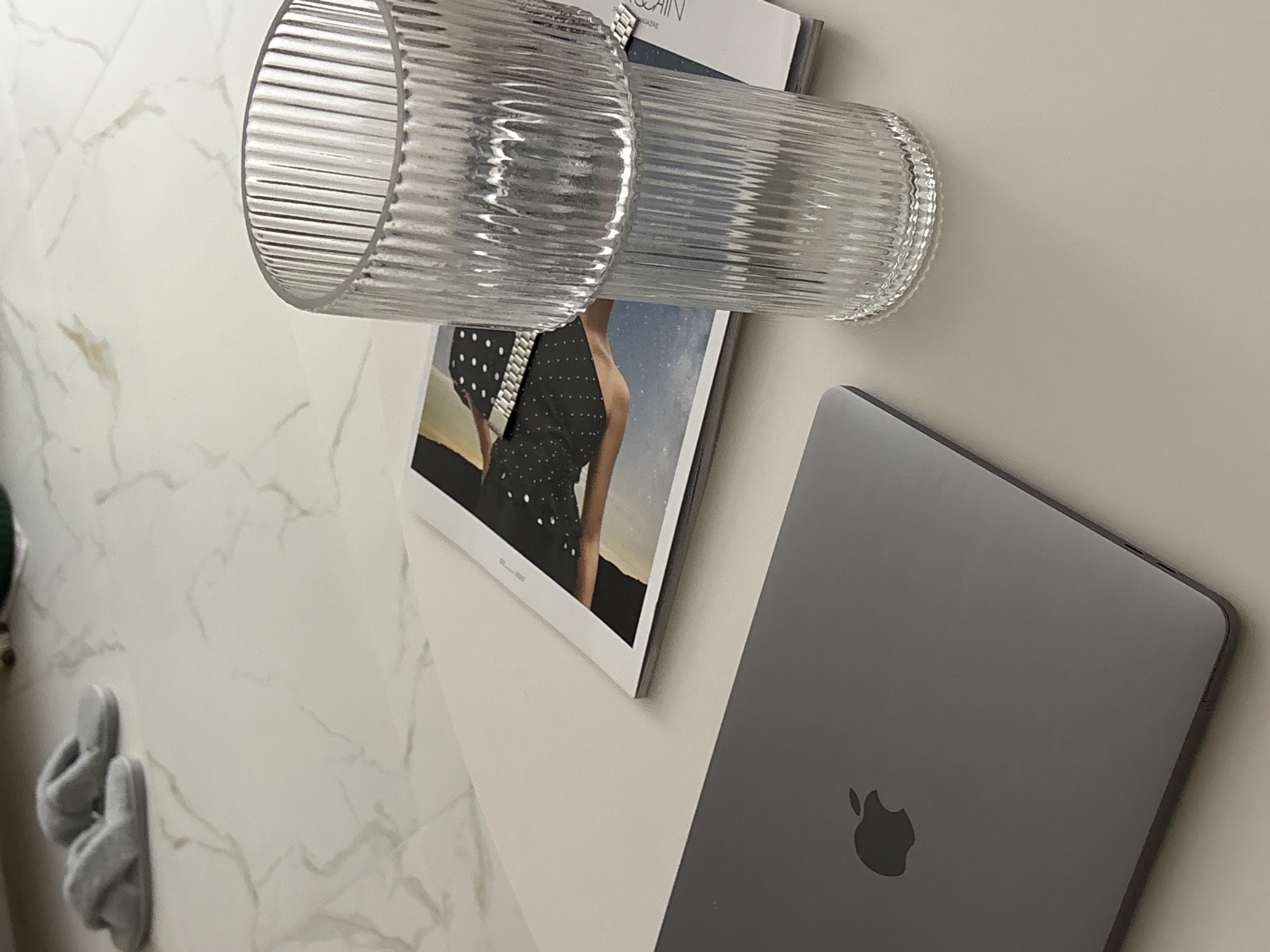 The image displays an overhead or high-angle view of a collection of items arranged on a flat, light-colored surface, likely a table or desk. The surface features a white marble-like pattern with grey veins on the left, transitioning to a plain off-white area on the right.
In the upper center, a clear, horizontally-oriented glass or vase with a distinct fluted or ribbed texture is visible. Directly below it lies an open magazine or book, showcasing a partial image on its page. This image depicts the torso of a person wearing a dark, possibly polka-dotted top. Above this image, the word "AGAIN" is clearly visible on the magazine.
To the right, a closed space gray Apple MacBook is positioned, its lid featuring the iconic Apple logo. In the bottom-left corner, resting on the marble-patterned surface, is a pair of soft, light grey slippers.
The scene is set indoors, with soft, even lighting that suggests either diffused natural light or artificial illumination. The arrangement of a laptop, magazine, glass, and slippers implies a relaxed or focused activity, such as reading, working, or simply unwinding at home. There are no people visible in the image.