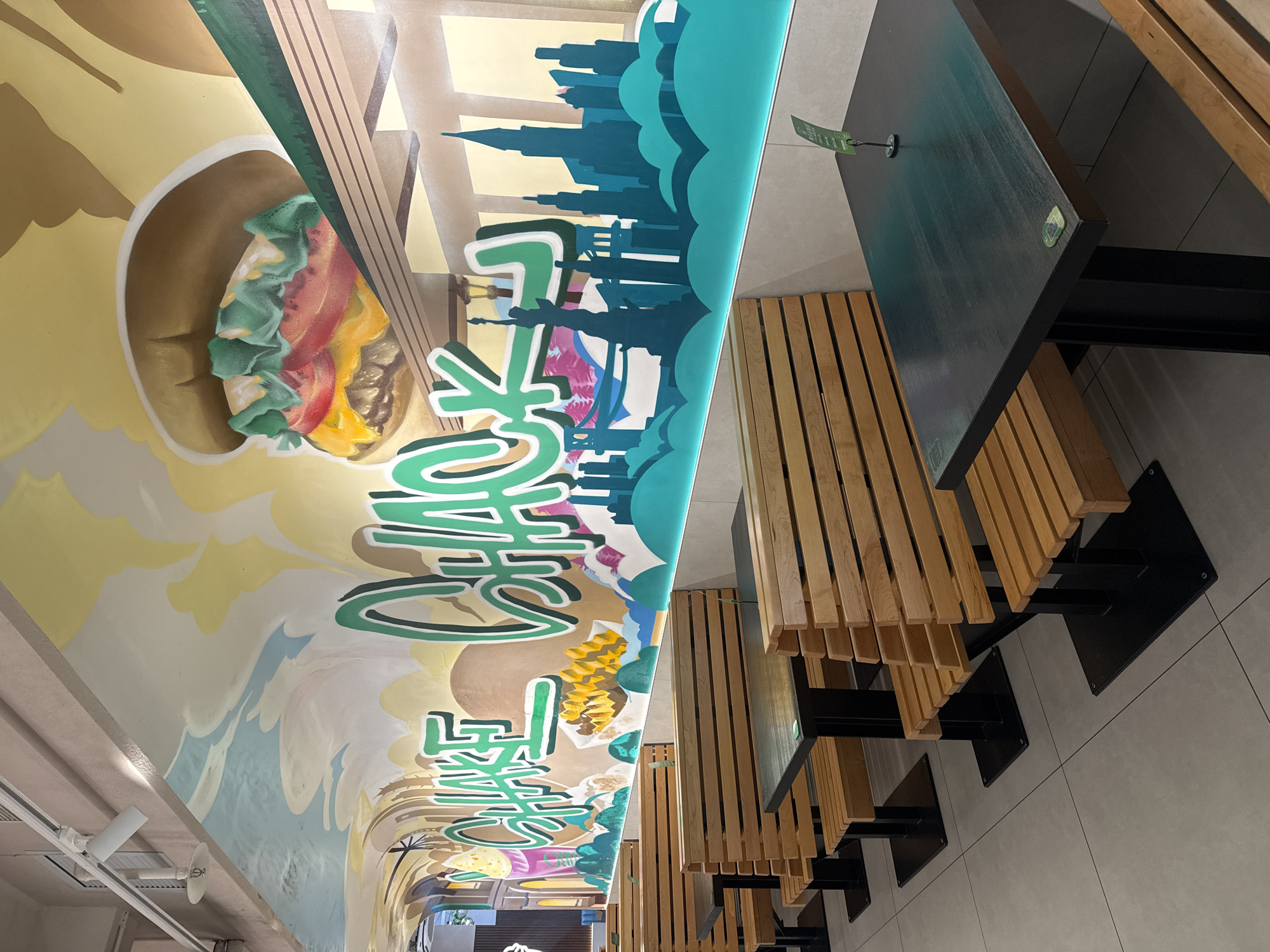 This is an indoor, eye-level shot of a Shake Shack restaurant in Shanghai, China. The walls and ceiling are adorned with a vibrant mural depicting a burger, fries, a milkshake, and the city skyline. Wooden booths with matching benches are arranged around dark tables. A green menu holder with a small green card is visible on one of the tables. The floor is tiled in a neutral gray. No people are visible in the image.