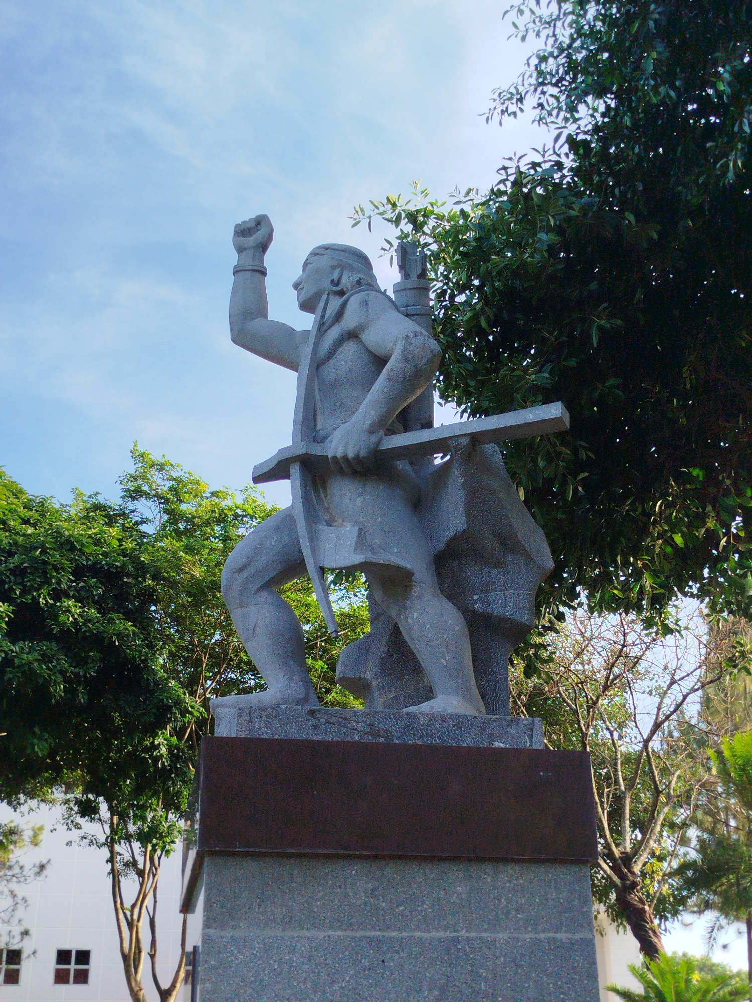 The image shows a stone statue of a man with a raised fist. He is holding a horizontal bar across his shoulders. The statue is mounted on a two-tiered base. The surrounding environment includes trees and a building in the background. The location is Pleiku Ward, Vietnam.