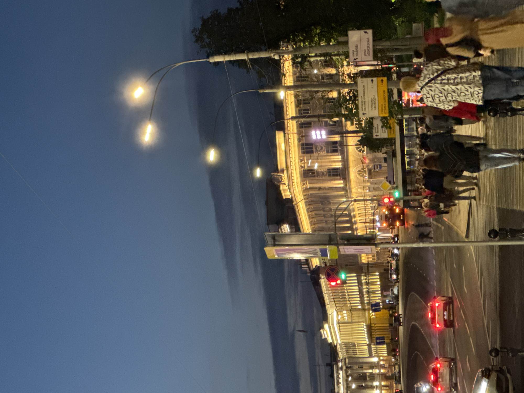 The image depicts a street scene at dusk in Nizhny Novgorod, Russia, likely on Bolshaya Pokrovskaya Street, a popular pedestrian thoroughfare. The sky is a deep blue with wisps of clouds. Ornate, illuminated buildings with classical architectural features line the street on the left and in the background. Streetlights cast a warm glow on the scene, with two prominent lampposts with multiple lights visible. Several cars are driving on the multi-lane road in the foreground, their headlights and taillights illuminated. Pedestrians are walking along the sidewalk, some in groups, dressed in casual attire. Signposts with Russian text are visible, indicating street names and directions. The overall atmosphere is lively and urban.