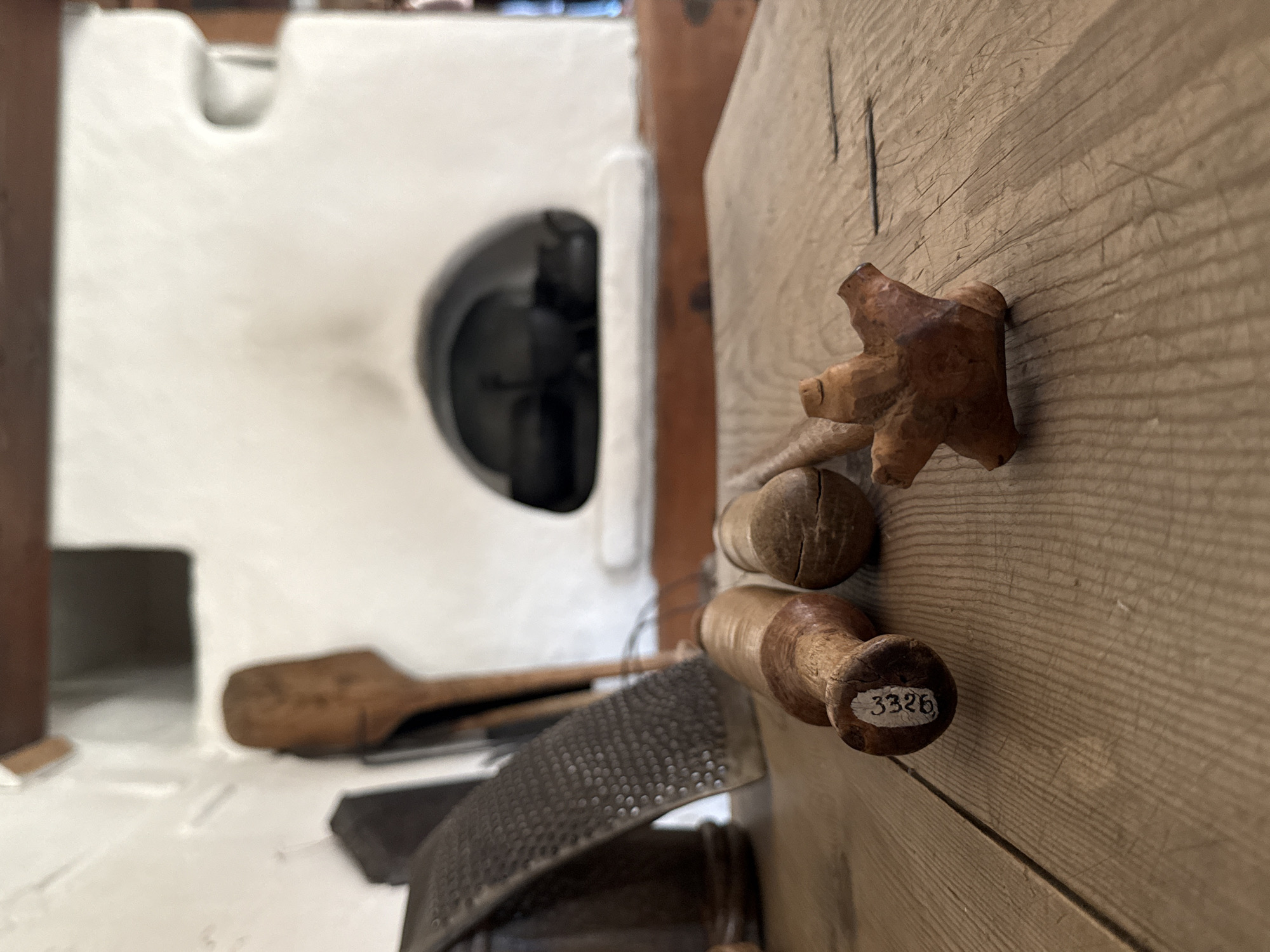 The image depicts a close-up view of wooden objects on a rustic wooden table in what appears to be a historical setting. In the foreground, three wooden tools are arranged on the table. The closest object on the left is a cylindrical tool with a lighter-colored wooden handle and a darker tip, bearing a white label with the number "3326". To its right is another cylindrical wooden object, possibly a pestle, with a cracked surface. Further to the right is a small, intricately carved wooden object resembling a star or a flower with multiple points.
In the background, a traditional stove, likely a Russian stove (pech), is visible. The stove is white-washed and has a dark arched opening where cooking pots are placed. To the left of the stove, a grater and another wooden utensil, possibly a spatula, are leaning against the wall. The environment suggests a historical dwelling or museum exhibit, potentially related to traditional Russian life and cuisine, likely located in Shushenskoye, Russia, a region known for its historical significance, particularly in connection with Vladimir Lenin's exile. The lighting is soft, and the focus is on the foreground objects, with the background slightly blurred, creating a sense of depth.