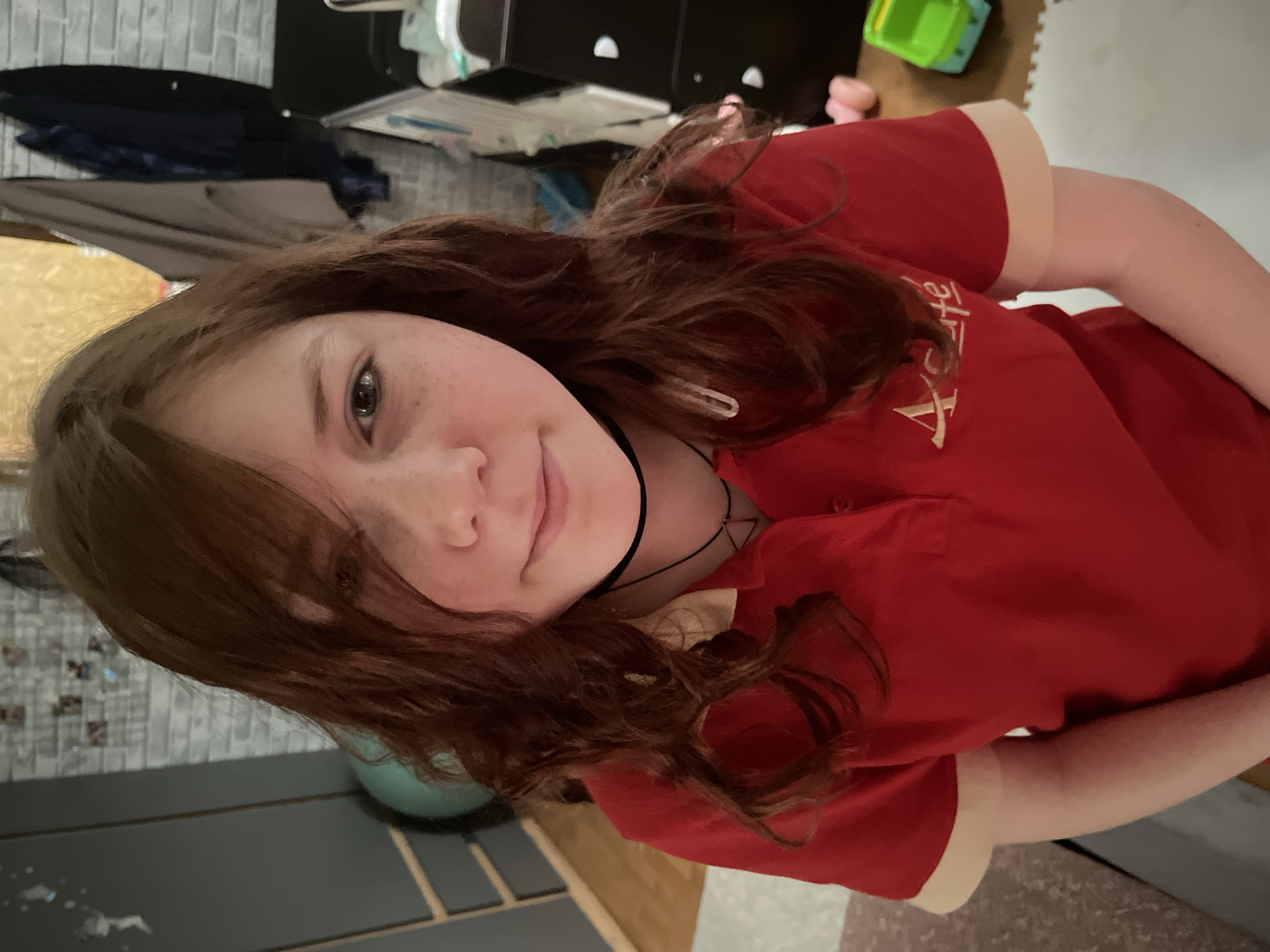 A young girl with auburn hair and freckles is facing the camera, wearing a red short-sleeved shirt with a beige collar and gold lettering. She has a black choker necklace. The background is somewhat cluttered, with what appears to be a gray brick-patterned wall, a black dresser, a green toy, and a foam floor mat visible. The location is unknown and the image doesn't specify that the picture was taken in the city of Vesyoloye, Russia.