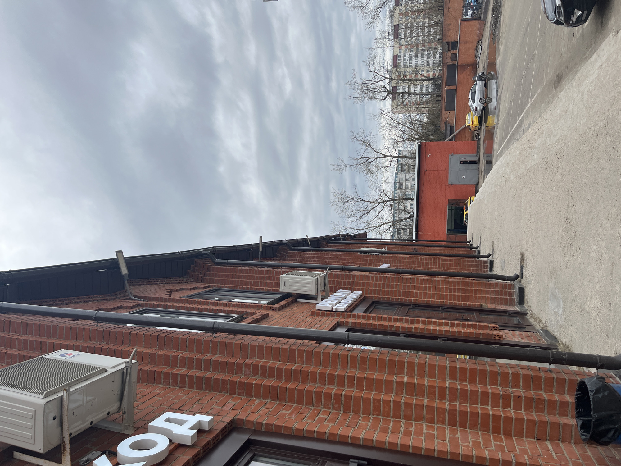 The image shows a brick building in Klin, Russia. The building has a dark roof and black pipes running along the side. There are several windows and air conditioning units mounted on the brick facade. White block letters are visible on the lower part of the building, appearing to spell out words in Russian. A grey overcast sky is visible above the building. In the background, there are taller residential buildings and a paved area with parked cars.
