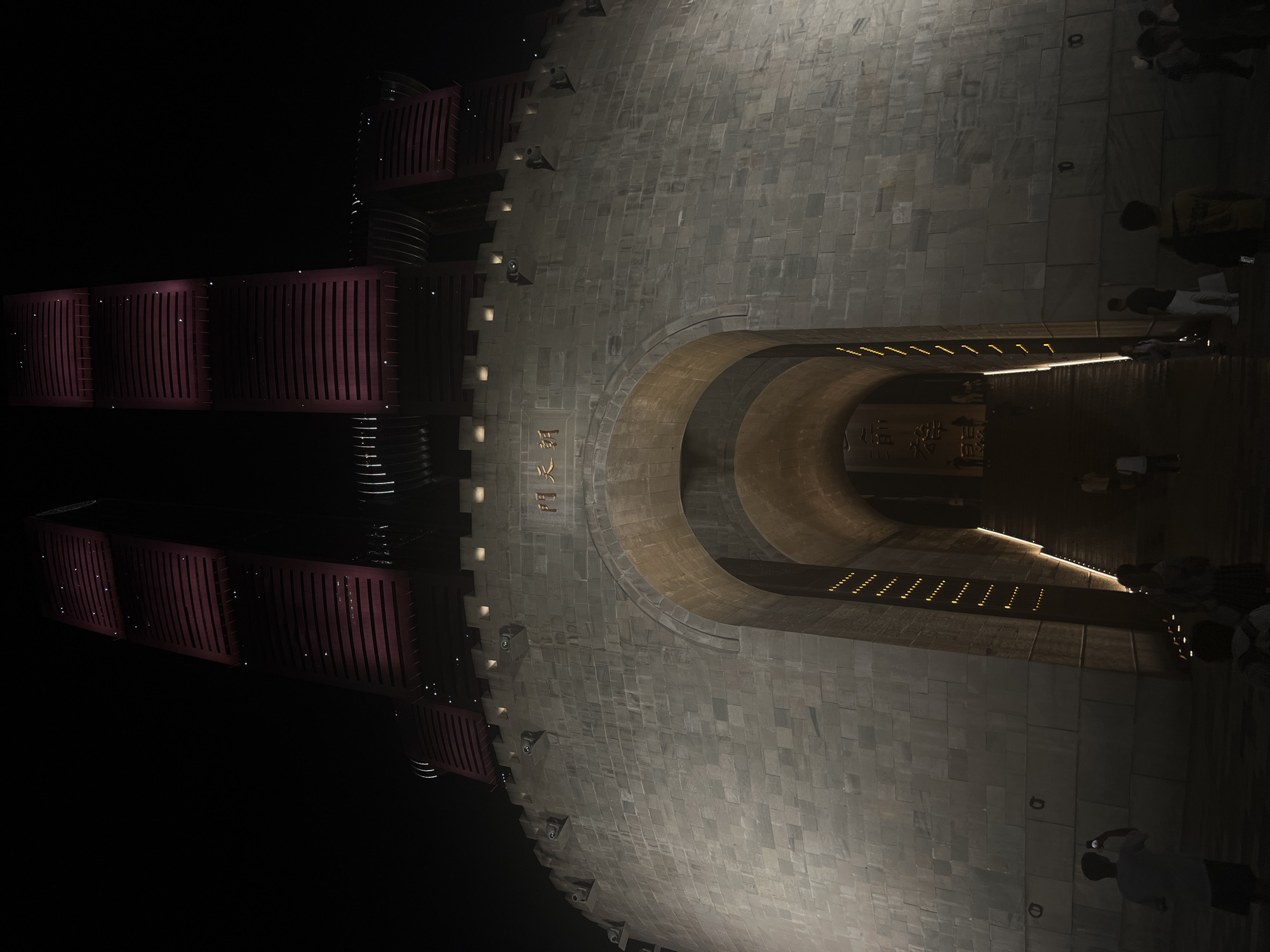 The image shows the exterior of the Hongya Cave building complex in Chongqing, Yuzhong District, China, at night. The building has a distinctive facade resembling ancient Chinese architecture, with a large arched entrance leading to a staircase. The upper levels of the building are illuminated with red lights, creating a striking contrast against the dark sky. Several people are visible in the foreground, some taking photos and others walking through the area. The stone walls of the lower part of the structure are lit, highlighting the intricate stonework. The scene suggests a bustling tourist attraction, even at night.
