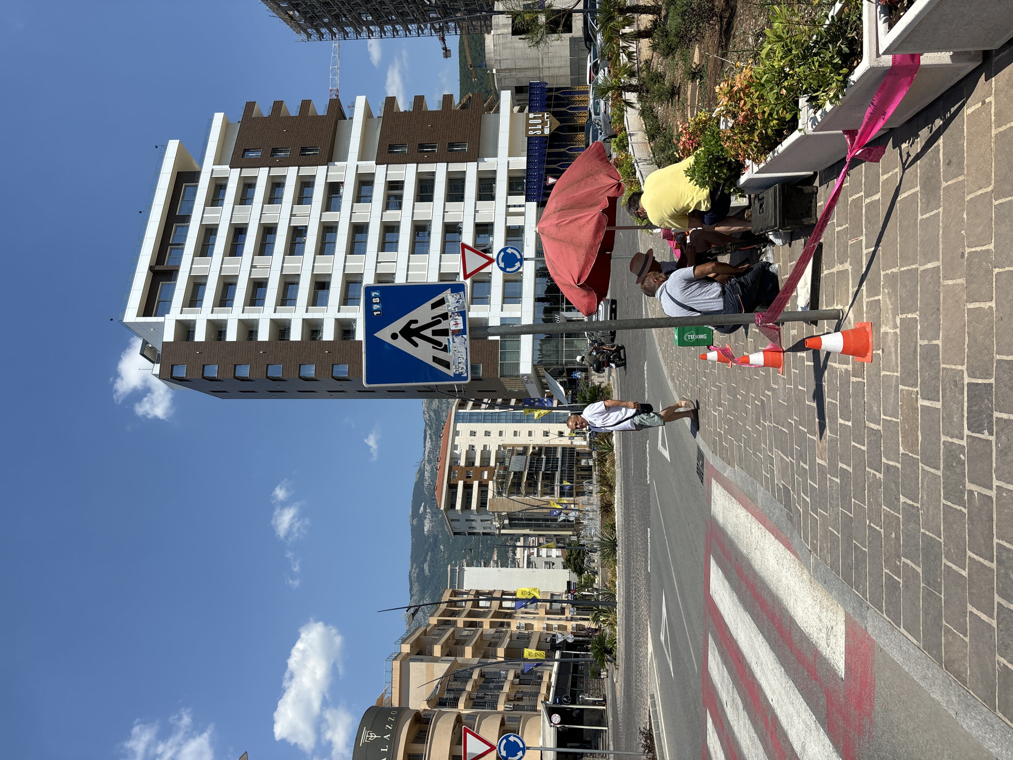 A bright, sunny day is depicted, with a clear blue sky and scattered white clouds. The scene is a street in Budva, Montenegro, with modern buildings lining one side of the road and a pedestrian crossing in the foreground.
Several people are present. In the mid-ground, a group of individuals are gathered near a red umbrella. One person in a yellow shirt appears to be interacting with equipment, possibly for a performance or event. Another person wearing a hat is also in this group. Further down the street, a man in a white t-shirt and shorts stands on the sidewalk, looking towards the camera.
Objects in the scene include traffic signs, such as a pedestrian crossing sign and a roundabout sign. Orange and white traffic cones are visible along the sidewalk, and pink caution tape is strung between planters. A dark speaker box is positioned on the ground near the people under the umbrella. The buildings are modern, with many windows and balconies. A mountain range is visible in the distance.
The overall atmosphere seems casual and relaxed, suggesting a typical day in a coastal town. The time of day is likely midday, given the strong sunlight and shadows. The presence of the cones and tape might indicate a temporary setup or restriction on the sidewalk.