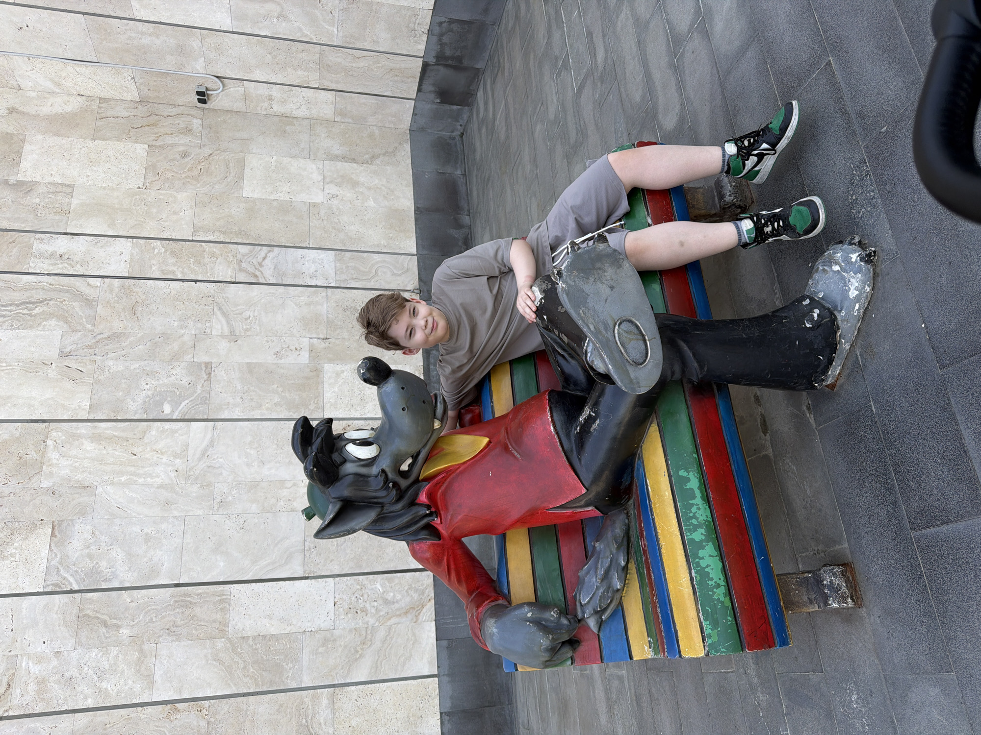 A boy is lying on the lap of a cartoon wolf bench statue located in Moscow, Russia. The boy has light brown hair and is wearing a grey short sleeve shirt and grey shorts. He has green and black sneakers on his feet. The statue is of a cartoon wolf. The wolf is wearing a red shirt with a yellow collar. The bench is painted in multiple colors, including blue, yellow, green, and red. The background consists of light-colored brick and dark grey pavement.