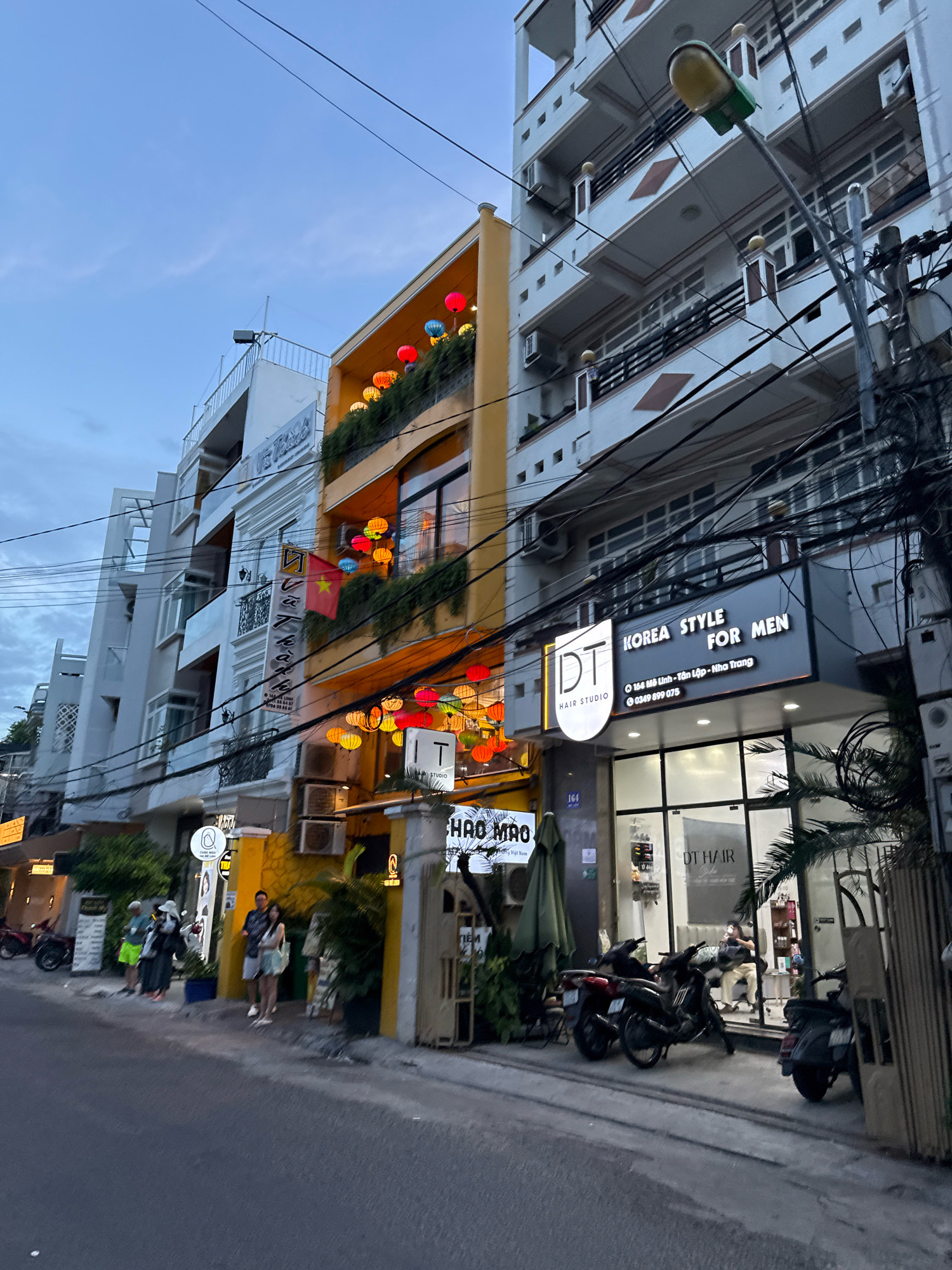 This image depicts a street scene in Nha Trang, Vietnam, in the late afternoon or early evening. The foreground shows a paved street and sidewalk. On the sidewalk, there are several parked motorcycles, including a dark-colored one in the center and a lighter-colored scooter on the right.
The buildings lining the street are a mix of styles. To the right, a white multi-story building with balconies and air conditioning units is visible, interspersed with a prominent lamppost. Adjacent to it, a bright yellow building features an upper floor adorned with colorful, illuminated paper lanterns. On this yellow building, a Vietnamese flag is displayed. Below, a business with a prominent sign reading "DT HAIR STUDIO KOREA STYLE FOR MEN" is visible, with large glass windows revealing the interior of a salon. Another sign below it reads "DT HAIR".
Further down the street, more multi-story buildings continue, some appearing white and modern, others with more traditional Vietnamese architectural elements. A few people are standing on the sidewalk, some appearing to be waiting or walking. One group of three individuals, one wearing a white hat, is standing near a doorway. Another couple, a man and a woman, are also on the sidewalk.
The sky above is overcast with clouds. The overall impression is a bustling urban street with commercial establishments and residential buildings. The presence of numerous electrical wires indicates a dense urban environment. The lighting suggests it is twilight, with artificial lights from the buildings and lanterns beginning to illuminate the scene.