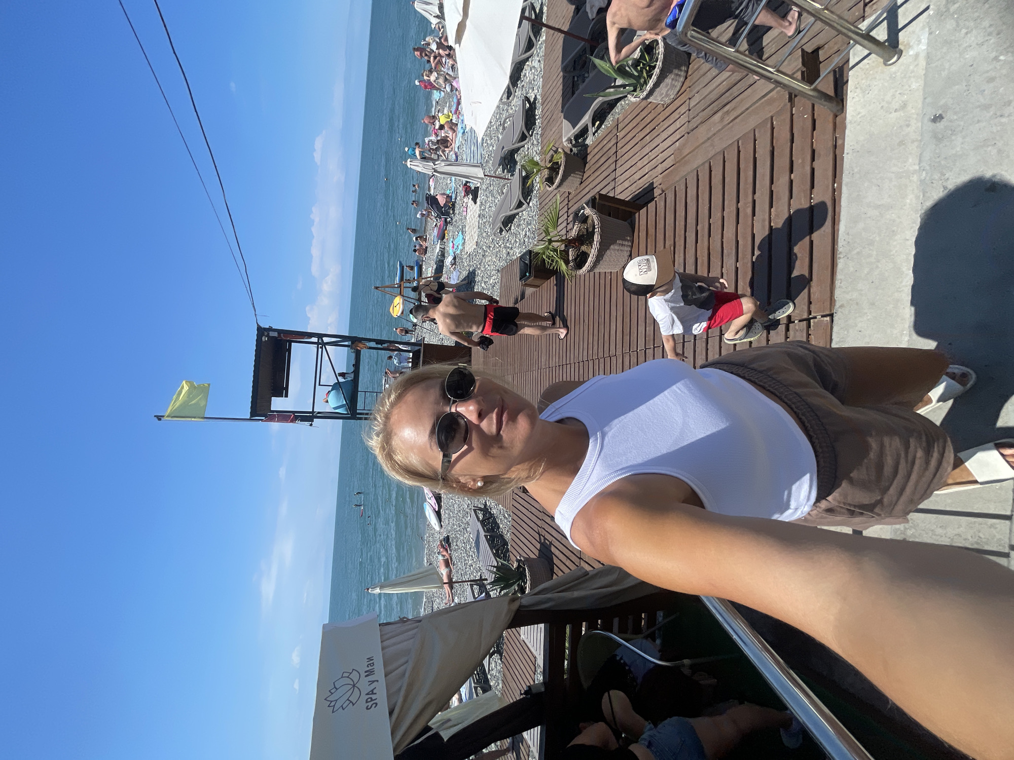 A candid, selfie-style photograph captures a blonde woman in sunglasses and a white tank top at a beach in Sochi, Russia. The woman is positioned in the foreground, with the beach and the Black Sea extending behind her. To her left, a canopy with the text "SPA у Маи" is visible. In the middle ground, a wooden walkway leads to a pebble beach dotted with lounge chairs, umbrellas, and numerous beachgoers swimming and sunbathing. A lifeguard tower with a yellow flag stands on the left side of the frame. A young boy in a baseball cap and red shorts is visible on the walkway in front of the woman. The sky is clear and blue, indicating a sunny day.