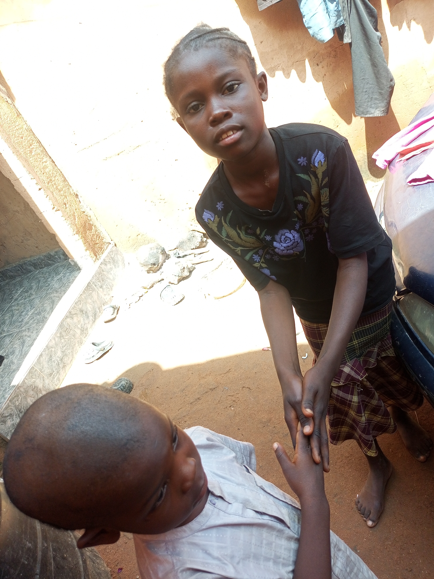 Two young children are interacting in an outdoor environment. A girl, with braided hair and wearing a black t-shirt with a floral design and a plaid skirt, is holding the hands of a younger boy. The boy, with a shaved head and wearing a light-colored, checkered shirt, is positioned below the girl, looking upwards. The ground appears to be dirt, with some scattered stones and a pair of sandals visible. In the background, there is a wall with some clothes hanging on it, and a vehicle is partially visible on the right side of the frame. The scene is lit by sunlight, casting shadows on the ground. The setting appears to be a residential area in a warm climate, possibly an informal settlement or courtyard.