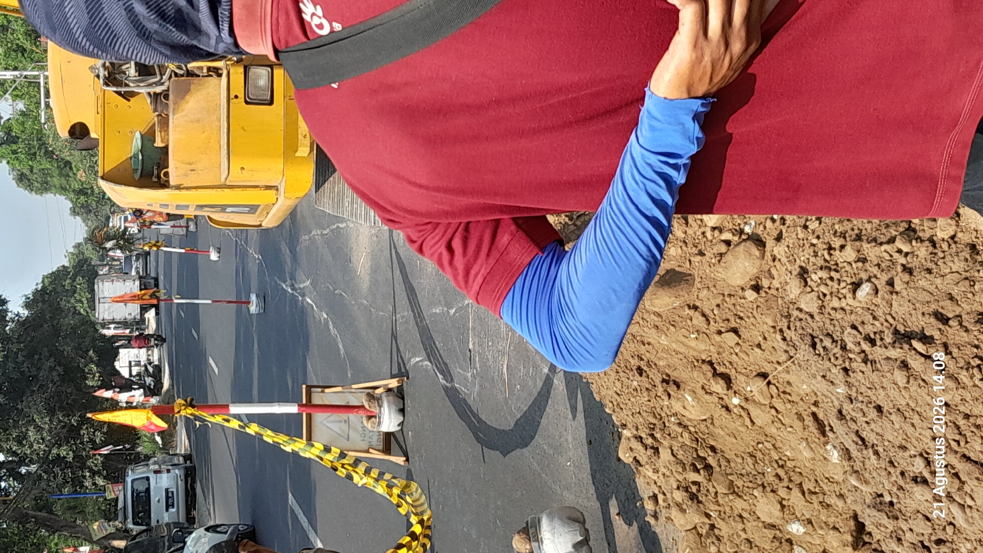 The image shows a section of a road, possibly under construction, in Takeran, Indonesia. In the foreground, a person wearing a red shirt and blue arm sleeves is standing, partially obscuring a pile of dirt. In the background, there is a yellow construction vehicle, traffic cones, and several vehicles including a silver van and a white truck. The scene appears to be during daylight hours. The date stamp "21 Agustus 2026 14.08" is visible in the lower left corner of the image, indicating the time and date the photograph was taken.