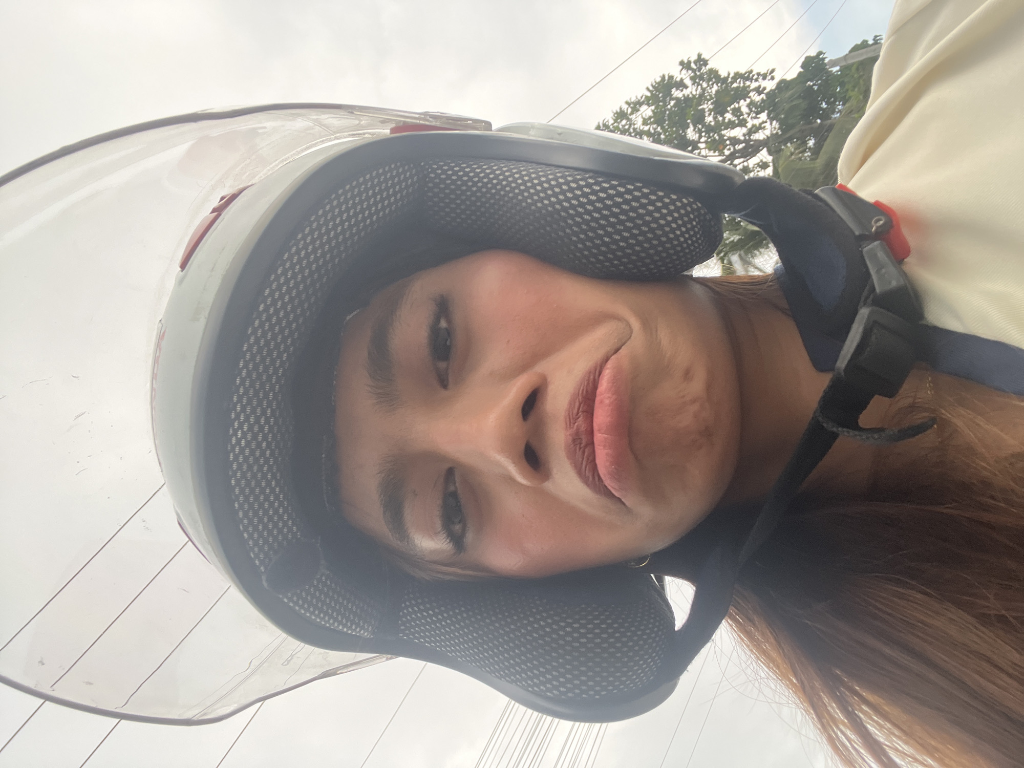 A person wearing a white and blue jacket and a white helmet with a clear visor is captured in a selfie. The helmet's clear visor is slightly smudged, and the person's face is tilted upwards, with their mouth pursed into a slight pout. Their eyes are open, and their eyebrows are slightly raised. The background shows a cloudy sky and some greenery, possibly trees, with electrical wires also visible. The person's long, brown hair extends down from under the helmet. The image appears to be taken outdoors, possibly while riding a motorcycle, given the helmet and the context of Davao City, Philippines. The time of day seems to be daytime, with overcast weather conditions suggested by the cloudy sky.