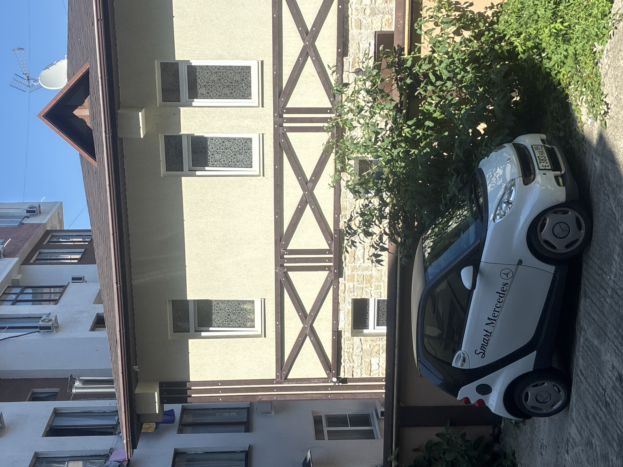 A white Smart car with black accents is parked on a paved surface in front of a light-colored building. The car has "Smart Mercedes" written on its side in black cursive font, along with the Mercedes-Benz logo. The license plate is visible and reads "E 383 HA 99". The building behind the car is multi-story with a tiled roof and several windows. Some of the windows have decorative patterned film. A section of the building features decorative dark brown wooden beams forming a criss-cross pattern over a stone facade. Lush green foliage is growing in front of the building and to the right of the car. The sky is clear and blue. This scene is in the city of Sochi, Russia, likely in a residential or commercial area.
