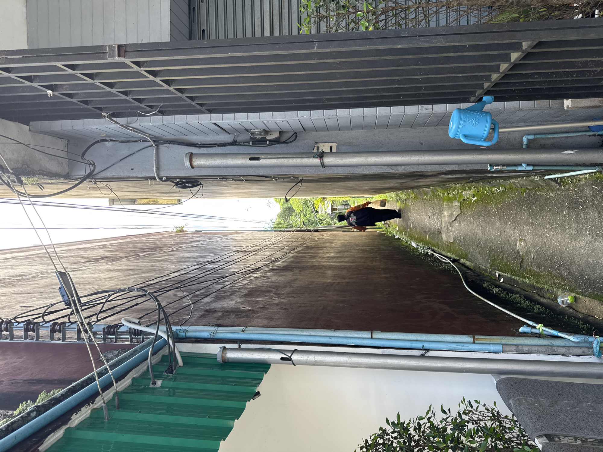 The image depicts a narrow alleyway in Ranong, Thailand. A person wearing a black jacket with orange accents and carrying a plastic bag walks away from the camera down the alley. The alley is flanked by two buildings; the one on the left has a dark brown, weathered wall, and the one on the right has a light gray brick facade with a black metal gate. Several electrical wires and pipes are visible running along the walls. Green foliage encroaches on the sides of the alleyway, and the ground is paved with concrete. A blue watering can is attached to a pole on the right side of the alley.