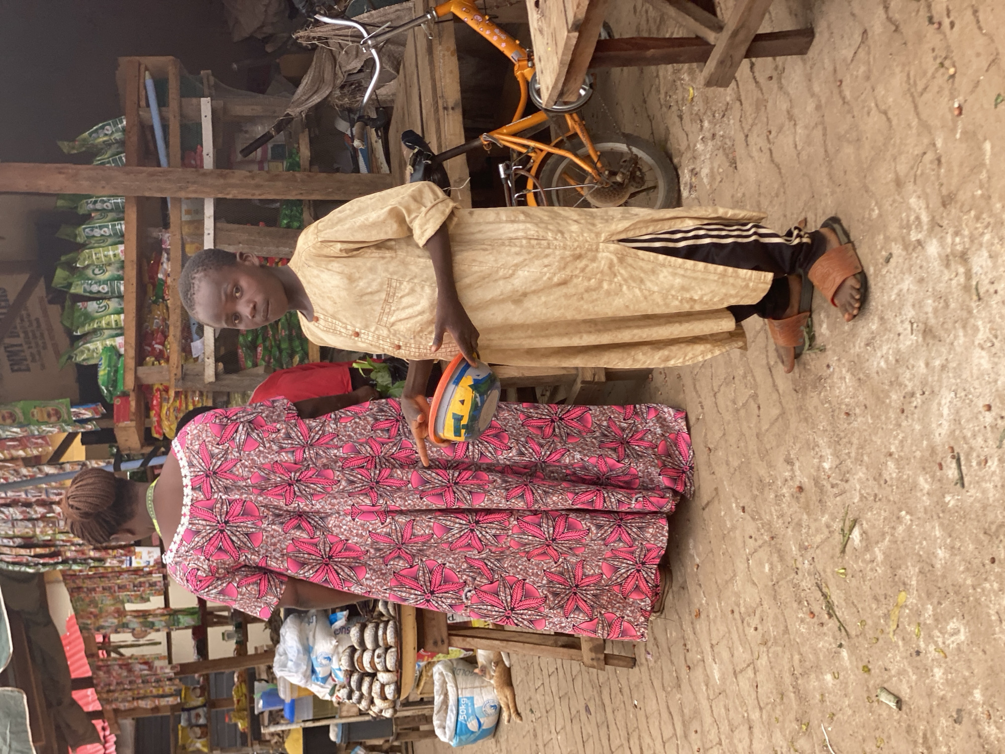 In a market setting in Kafanchan B, Nigeria, a young boy stands alongside a woman dressed in a pink floral patterned dress. The boy is wearing a light brown tunic and Adidas-striped pants and sandals. He holds a multi-colored bowl. The woman stands near a market stall laden with goods, including bags of flour and stacks of donut-shaped pastries. In the background, an orange bicycle is visible, along with wooden shelving holding various items. The ground is made of brick.