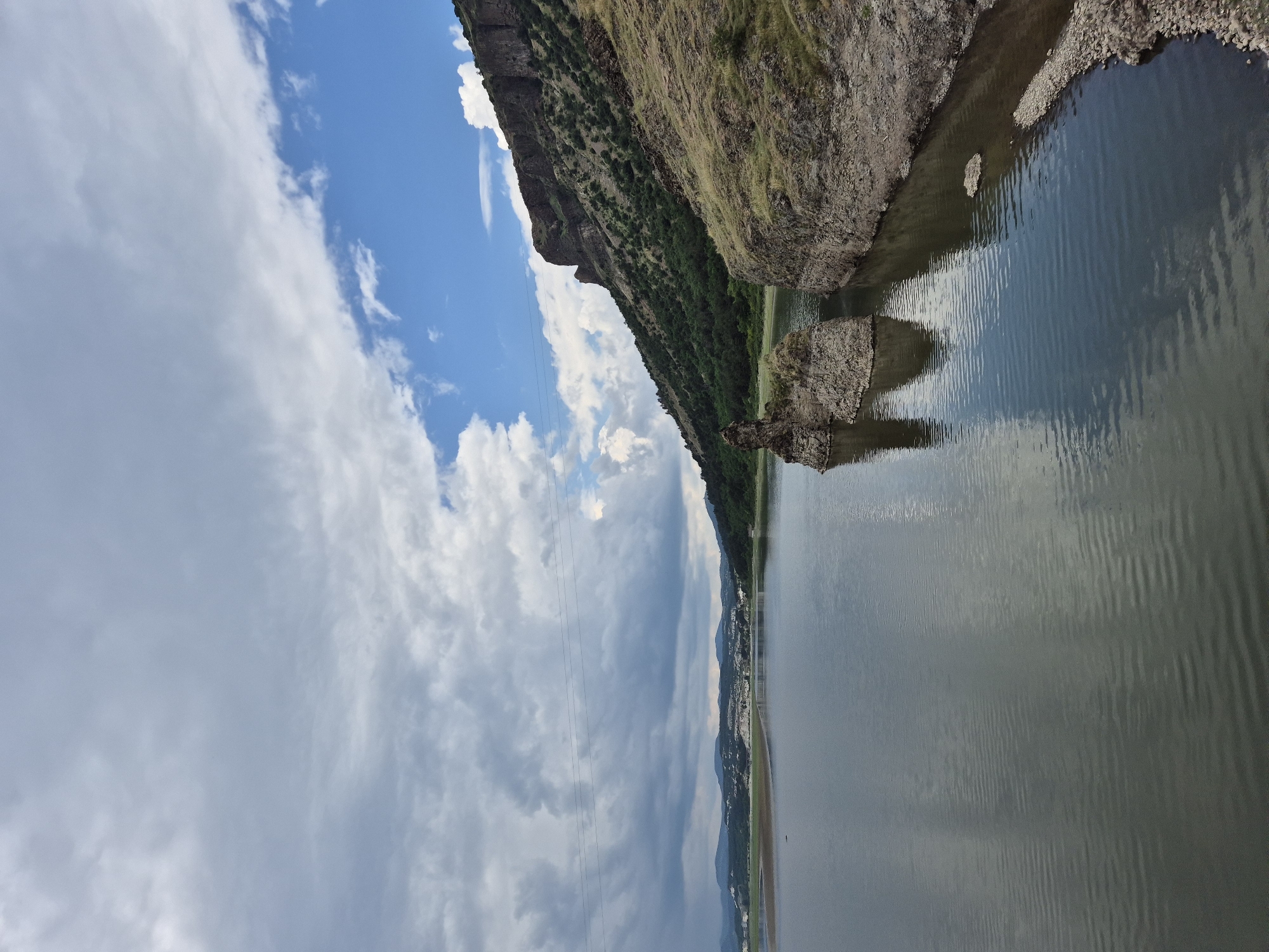 The image depicts a wide landscape scene in Lisitsite, Bulgaria, focusing on a large body of water bordered by mountainous terrain. The sky is split diagonally, with dense, grey overcast clouds dominating the upper left and clear blue sky with scattered white cumulus clouds in the upper right.
On the right side, a steep, rocky, and partially vegetated slope descends into the water. The rock formations are dark grey and rugged, covered sparsely with dry, yellowish-green grass and small green shrubs. Closer to the viewer, a rocky shoreline with small pebbles extends along the bank. Two distinct, dark, eroded rock formations, resembling stacks or small islets, rise from the water near the right bank, casting reflections.
The expansive body of water, likely a river or reservoir, occupies the lower and central portion of the frame. Its surface is calm with subtle ripples, reflecting both the grey clouds and the blue sky. The water color ranges from dark greyish-green to dark blue.
On the distant left bank, a sprawling settlement, identifiable as Lisitsite, is visible. It consists of numerous light-colored buildings nestled against lower hills and extending along the water's edge. Beyond the settlement, rolling hills or mountains with muted green foliage are visible under the overcast sky. Three distinct power lines stretch horizontally across the mid-upper section of the image, traversing the scene from left to right.
No people, animals, or specific actions are visible in the frame. The scene conveys a natural landscape with a distant human settlement under variable weather conditions.