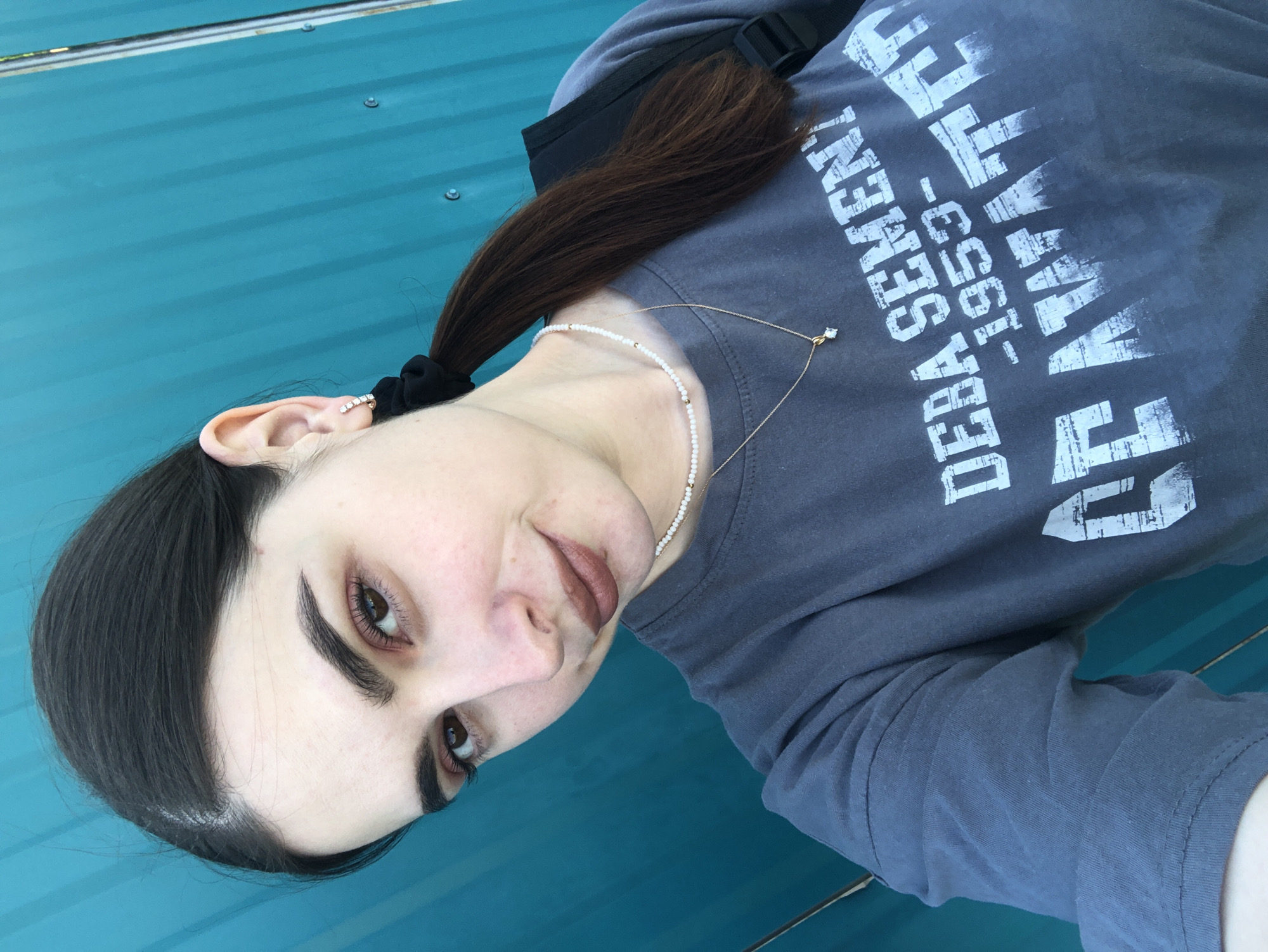 A selfie featuring a person with dark hair pulled back in a ponytail, wearing a gray t-shirt, and jewelry (earring, necklace). The t-shirt has white text, partially legible as "DEBA SEVEMENT 1953-". The background is a solid, teal-colored surface, potentially a corrugated metal structure. The location within Noviy, Russia, is not discernible from the provided image.