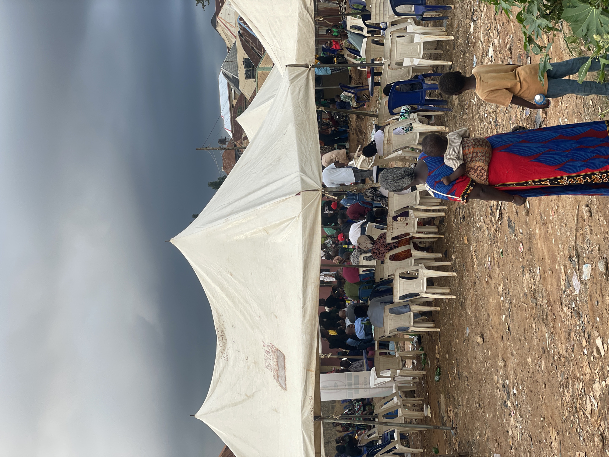 In Kurunduma, Nigeria, under a cloudy sky, a gathering of people are seated under large white tents. The ground is rough and uneven, covered with dirt, rocks, and scattered litter. Many white plastic chairs are arranged in rows, with some blue ones interspersed. In the foreground, a woman wearing a vibrant red and blue dress carries a baby on her back, secured with a patterned wrap. Beside her, a young boy in a tan shirt and blue jeans stands facing away from the camera, holding a bottle of water. In the background, beyond the tents, buildings with various roofing materials are visible. The scene suggests an outdoor community event or meeting.