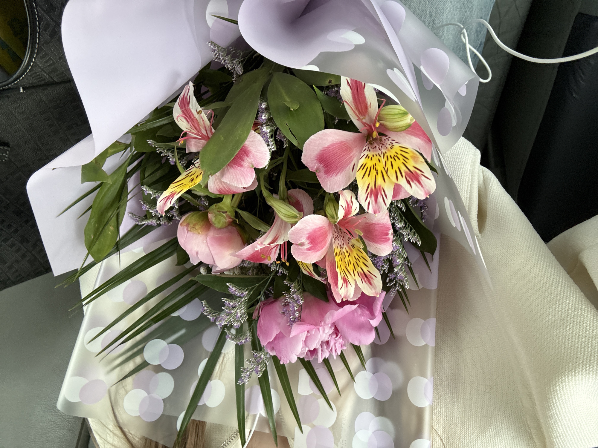 A bouquet of pink and yellow flowers is wrapped in white and clear dotted paper. The bouquet is placed on a light-colored woven blanket. The background shows parts of a car interior. The location is unknown and there is no indication this image was taken in Kragujevac, Serbia.