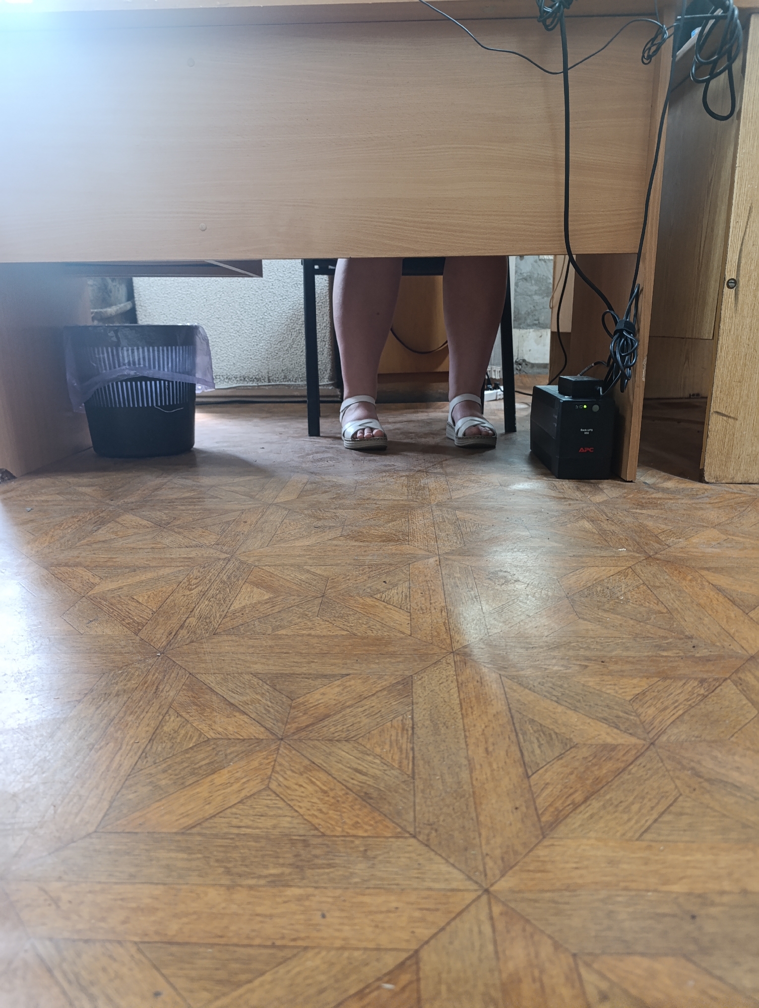 The image shows the legs of a person wearing white sandals, standing under a wooden desk. To the left of the person's legs is a black trash can with a plastic liner. To the right of the person's legs is a black APC uninterruptible power supply (UPS) unit with cables. The floor is made of wooden parquet flooring with a geometric pattern. The background behind the desk appears to be a wall with some pipes and a rough texture. The scene is dimly lit, with some light coming from above the desk.