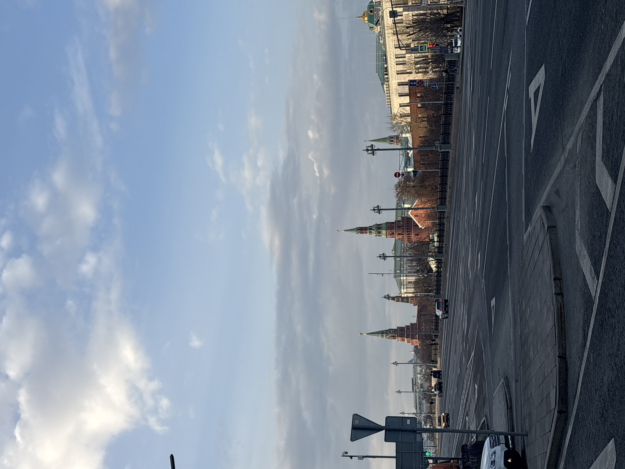 The image depicts the Moscow Kremlin from across the Moscow River. The view includes the towers and walls of the Kremlin, as well as nearby buildings and part of the city skyline. In the foreground, there is a road with markings. There are street lights along the road and some vehicles in the distance. The sky is partially cloudy.