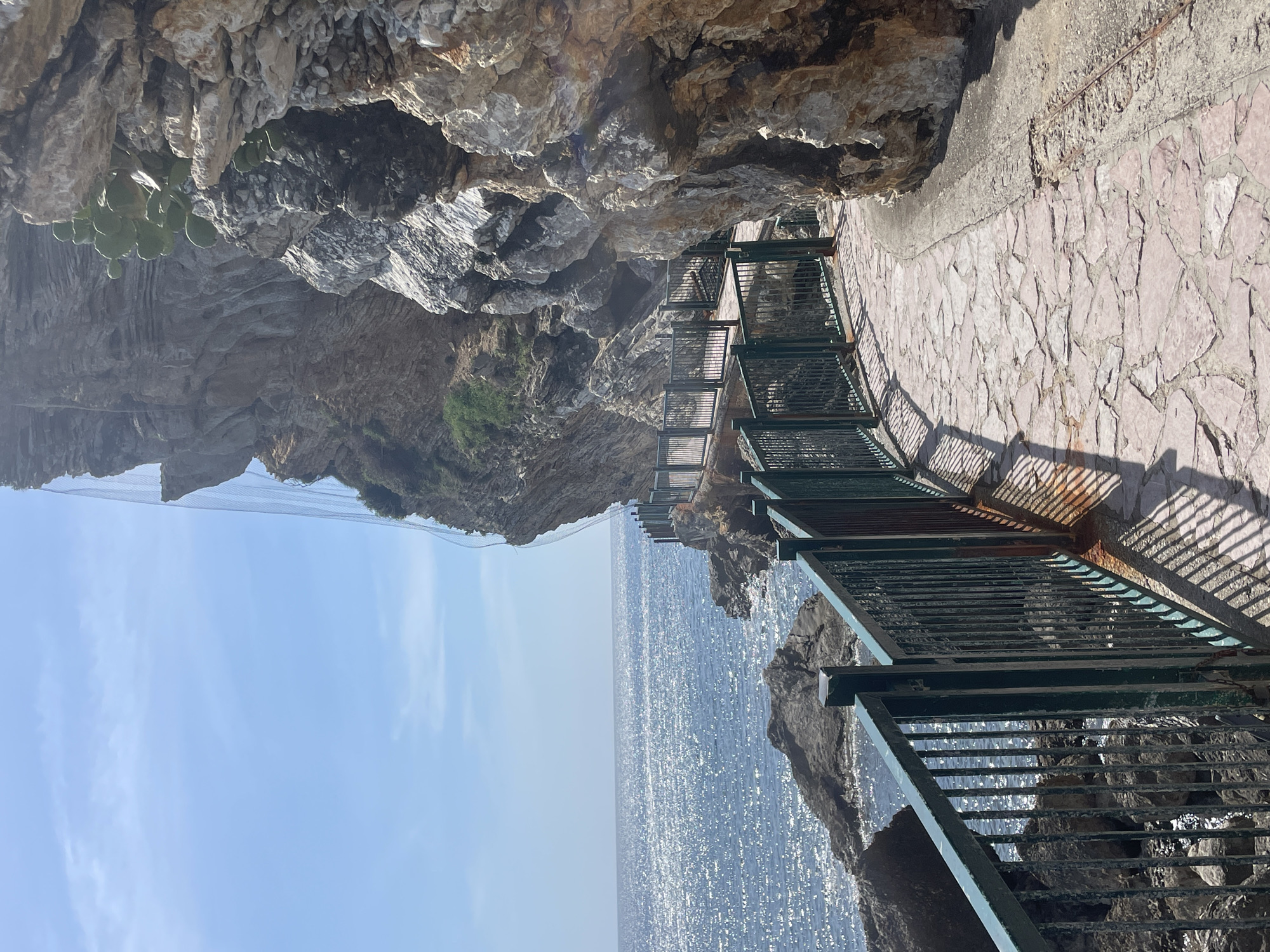 The image presents a sunny coastal landscape along the Budva Riviera, Budva, Montenegro, depicting a section of the pathway connecting Budva Old Town to Mogren Beach. A winding pedestrian path, constructed from irregular light-colored flagstones, dominates the foreground and midground, curving around a formidable rocky cliff face on the right. A dark green metal railing with vertical bars, showing signs of rust at its base, lines the path's seaward edge, casting sharp, elongated shadows onto the flagstones.
To the left, the expansive blue sea stretches horizontally, its surface shimmering intensely with sunlight. Dark, irregular rock formations are visible within the water near the coastline. Above, the sky is clear and light blue, with minimal, wispy cloud cover. The dominant cliff on the right comprises rugged, stratified rock in shades of grey and brown. Sparse green vegetation, including what appears to be a prickly pear cactus, clings to the cliff face. A fine-mesh safety net is partially visible against the upper portion of the cliff, suggesting rockfall protection measures. No individuals or active movements are present in the static scene.