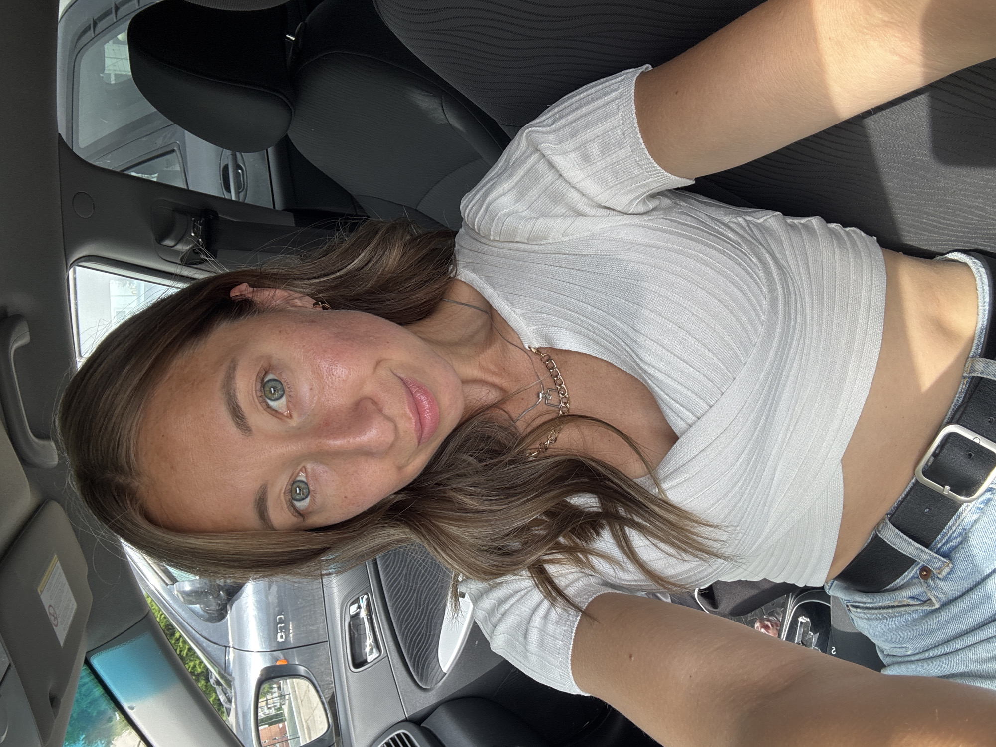This media file presents a close-up portrait of a young woman inside a car, taken in Chaikovsky, Russia. The woman, with long, wavy brown hair, fair skin, and striking blue-green eyes, is looking directly at the camera with a soft, gentle smile. She is wearing a fitted white ribbed crop top, light blue denim jeans, and a black belt with a silver buckle. A delicate gold chain necklace with a small, square-shaped pendant and a small gold hoop earring are visible accessories.
The setting is the interior of a vehicle, likely the front seat, captured during bright daytime hours. Strong sunlight casts noticeable shadows, particularly across her right arm and the car's dark upholstered seats, indicating a clear, sunny day. Through the windows, other vehicles can be seen, suggesting an urban or suburban street or parking lot. A gray pickup truck or SUV is partially visible through the side mirror, with the text 'RAM' clearly identifiable on its side. The car's interior features black and gray components, including the dashboard and seats. The overall impression is a casual, self-taken photograph in a relaxed environment.
