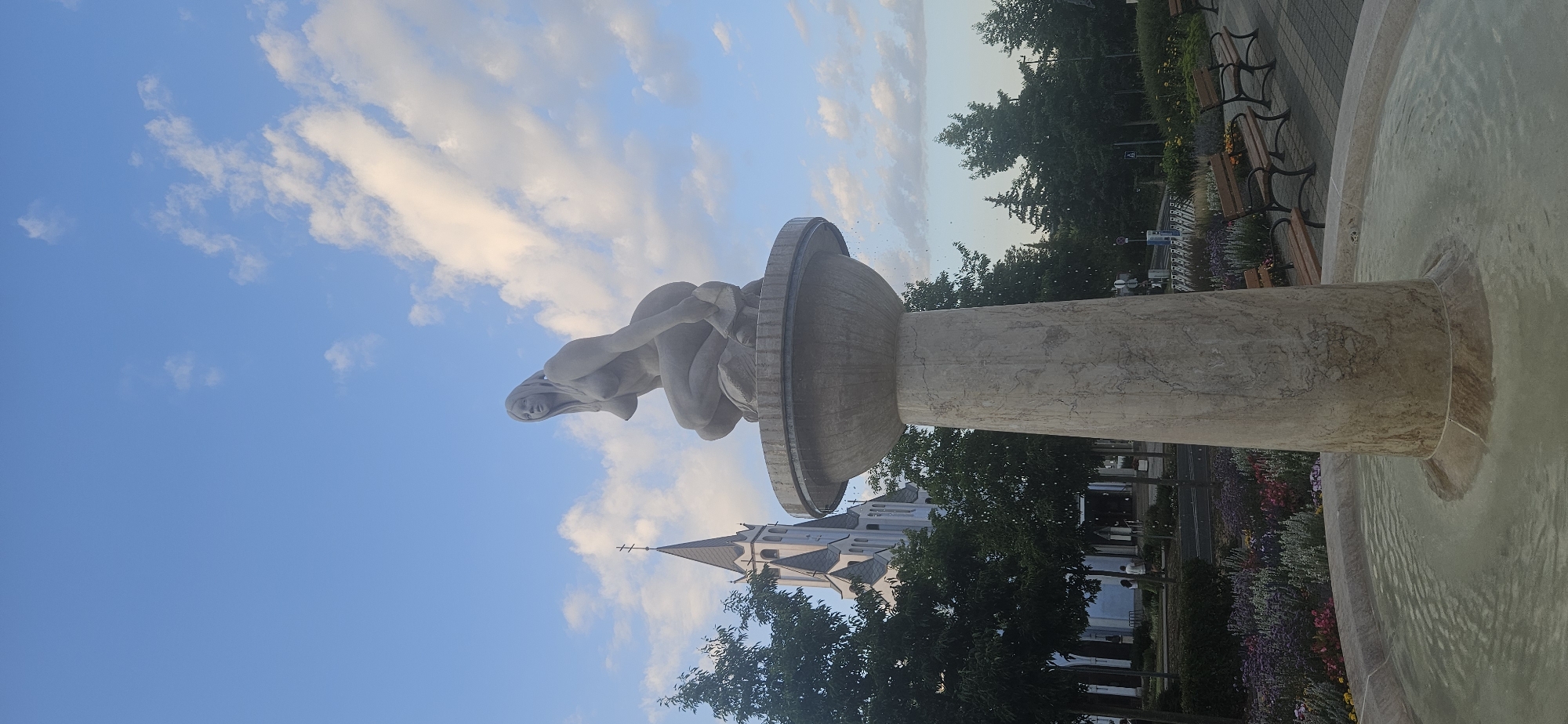 The image shows a statue of a woman in a fountain located in the city of Hévíz, Hungary. The statue is made of stone and depicts a nude woman kneeling. The fountain has a large stone pedestal. In the background, a church with a tall spire is visible, along with trees and a blue sky with white clouds. Benches and flower beds are also present in the foreground, suggesting a public park or square. The water in the fountain is calm, with ripples indicating it is active.