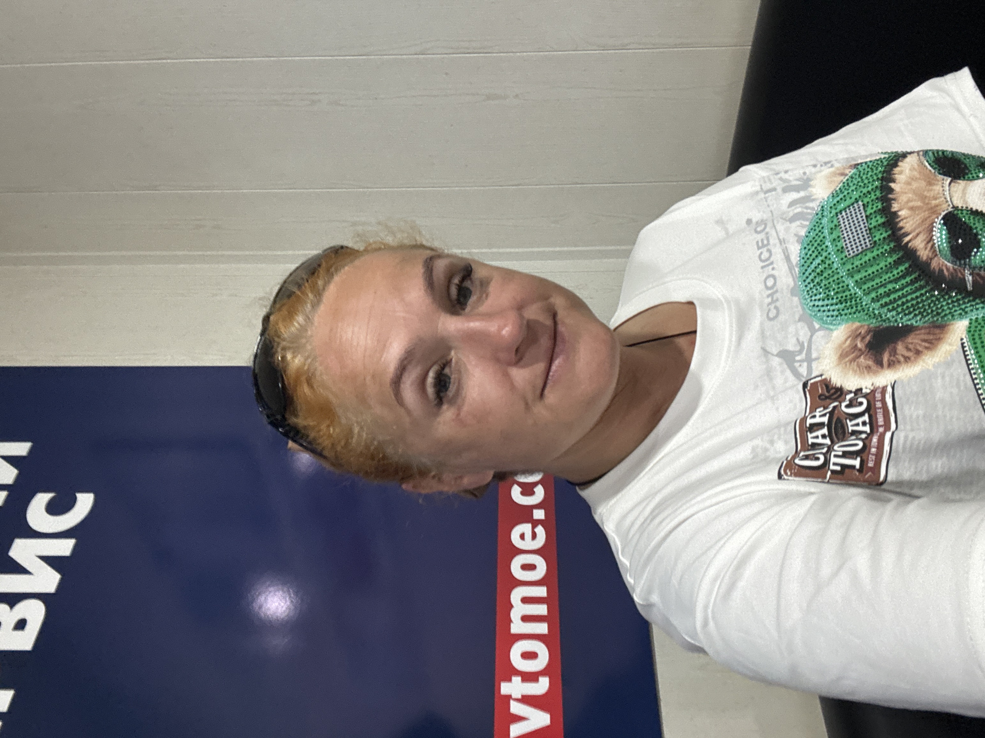 A woman with dyed blonde hair and sunglasses on her head is sitting in front of a blue banner. The banner has white Cyrillic text and a red horizontal stripe with white text that reads "vtomoe.co". The woman is wearing a white t-shirt with a graphic of a bear wearing a green hat and sunglasses. The background behind the woman is a light-colored, vertical paneled wall. The scene appears to be indoors, potentially in a shop or office, given the banner and wall paneling.