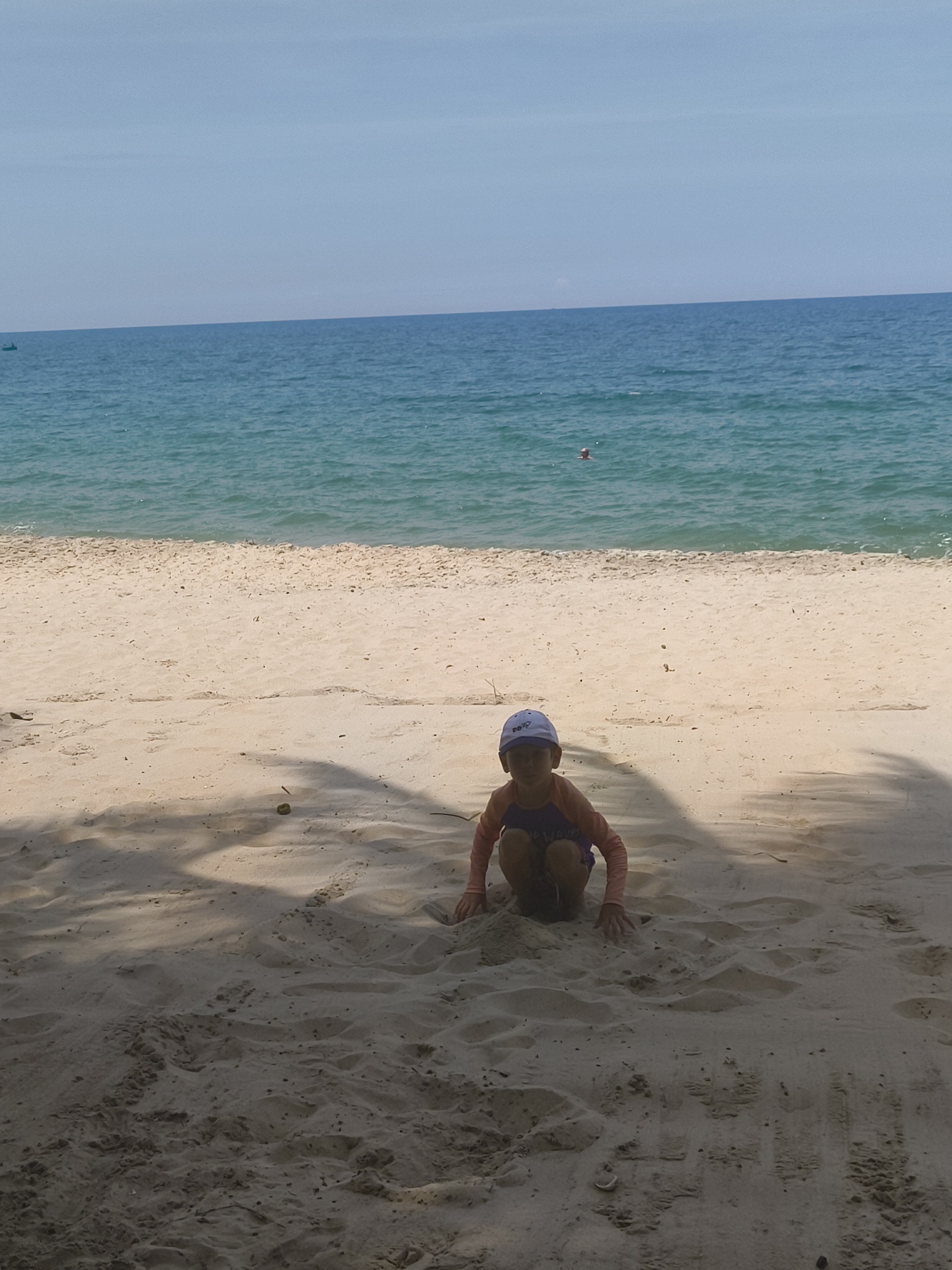 A child wearing a purple and orange rash guard and a blue cap squats on a sandy beach. The child is positioned in the foreground, partially in shadow. The ocean, with calm, blue water, is visible in the background, meeting the horizon under a pale sky. The beach is composed of light-colored sand with scattered debris and shadow patterns from overhead foliage. There is a distant boat on the water and another person swimming. The scene is devoid of urban elements and appears to be a natural coastal environment.